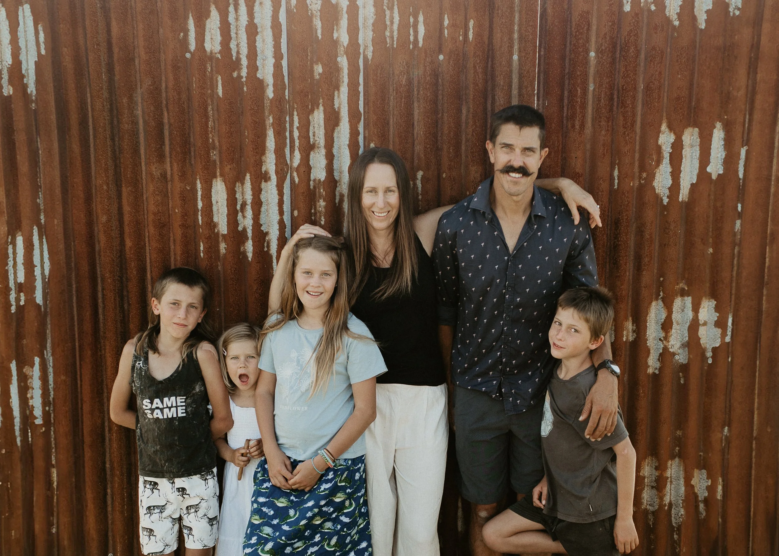 A group of six people, including children and adults, standing together in front of a rusted metal wall. They are smiling and looking at the camera, with some children having their arms around each other or adults.