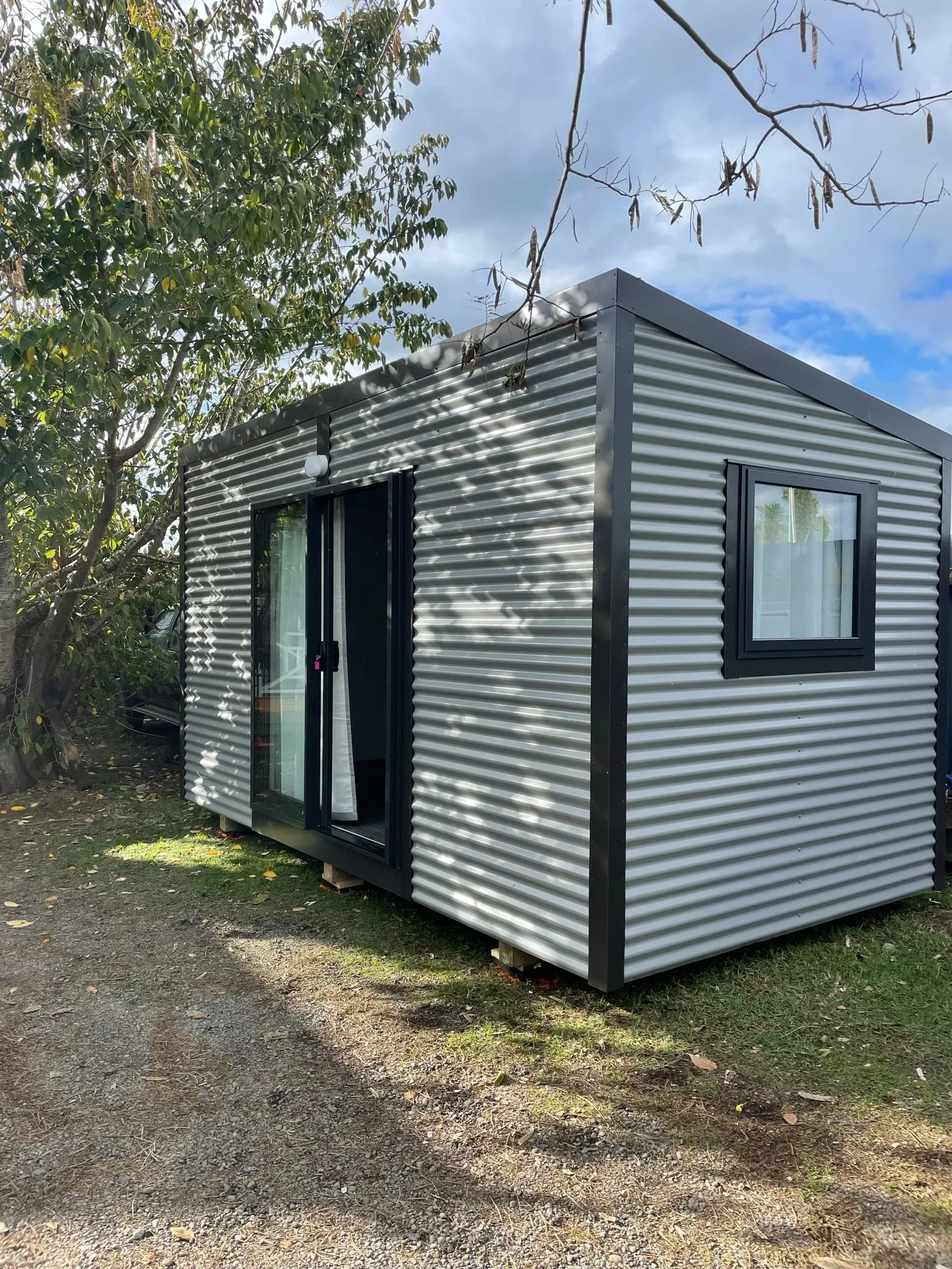 A small, modern tiny house with metal siding and black-framed windows, situated outdoors on a patch of grass and gravel with trees and blue sky in the background.