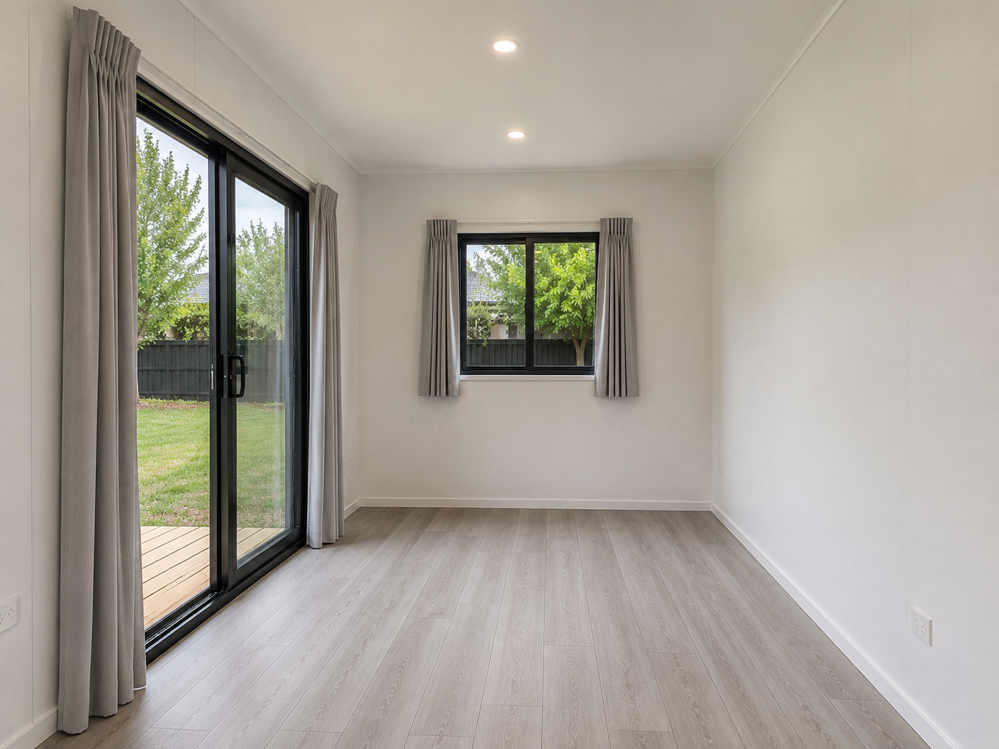 Empty room with light wood flooring, white walls, a sliding glass door with curtains leading outside, and a window with curtains showing a backyard with trees.