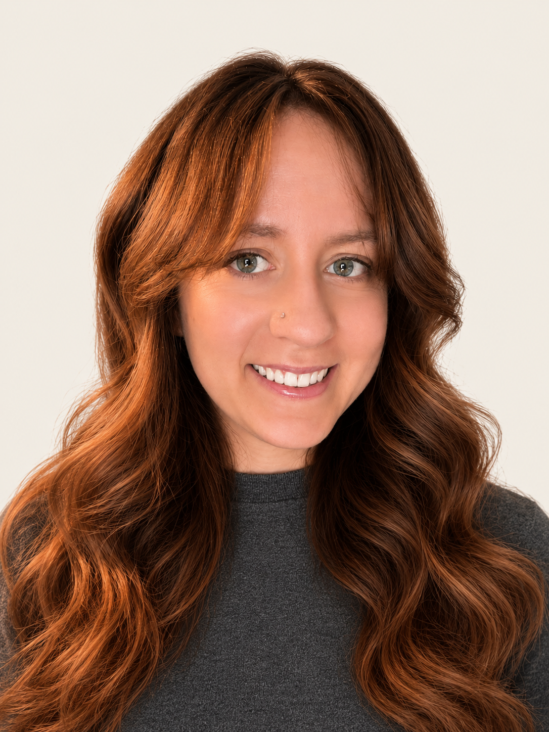 A smiling woman with long wavy red hair, wearing a dark gray shirt, against a plain off-white background.