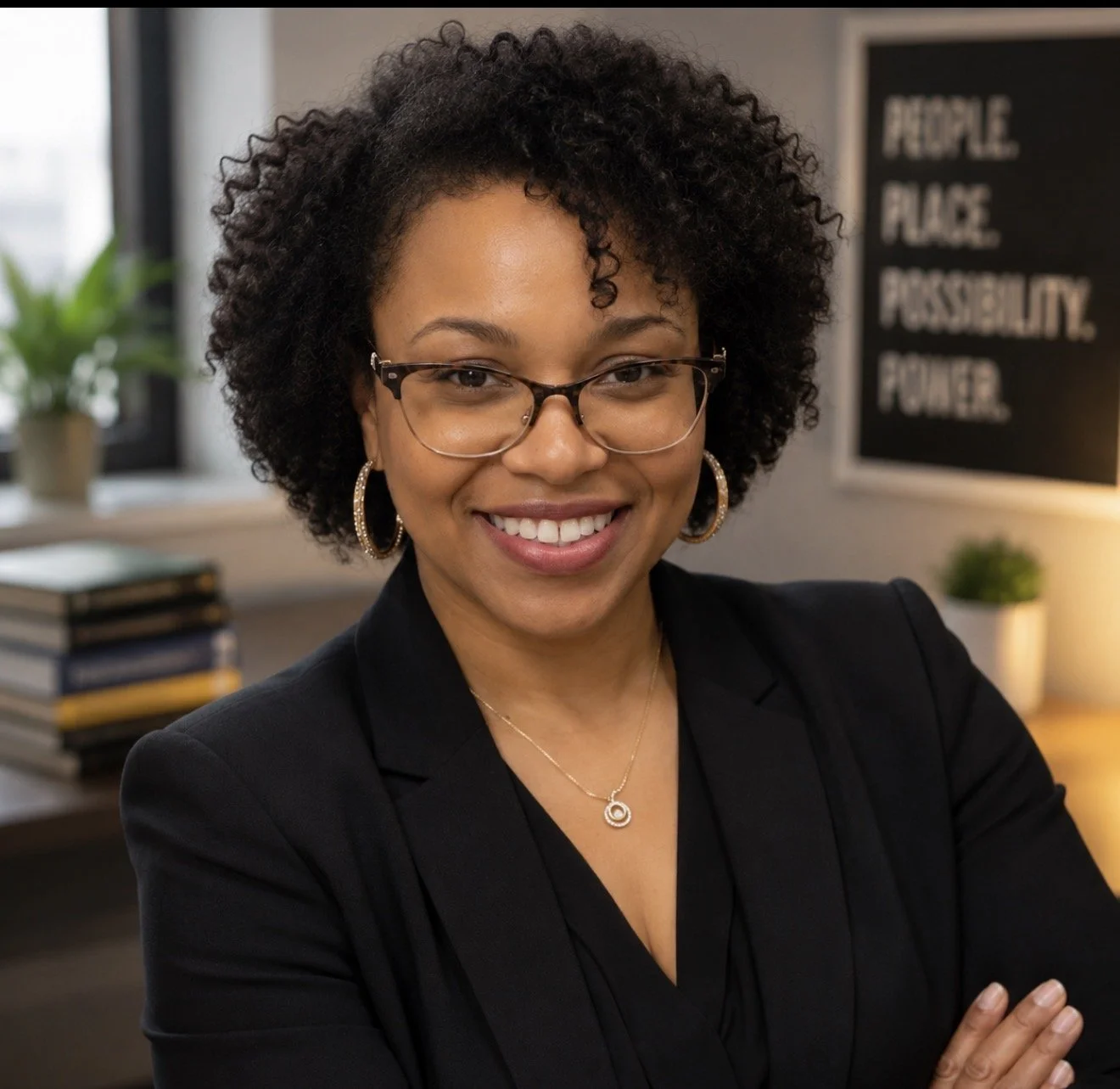 Founder of INS, that is a Black woman is Smiling woman with glasses and curly hair, wearing a black blazer and jewelry, standing in an office environment. There is a sign in the background that says People Place Power