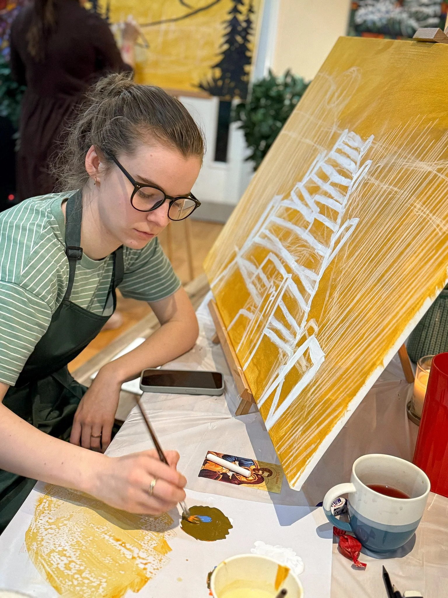 A woman with glasses and a bun hairstyle painting a picture on a canvas, the painting is in the early stages and the image is sketched on the canvas with chalk.