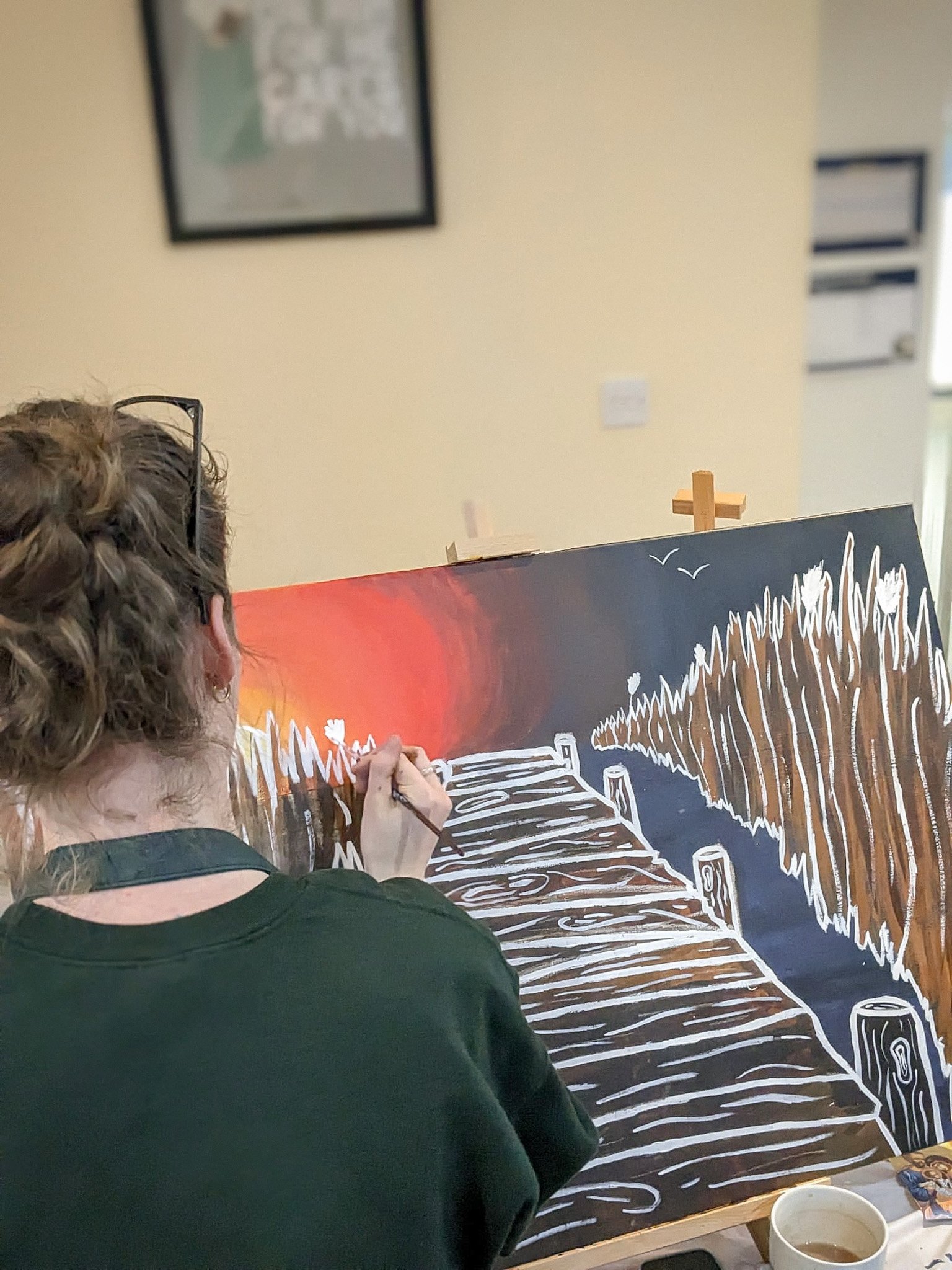 Person painting a blackboard scene of a wooden bridge over a creek with trees and birds with acrylic paints.