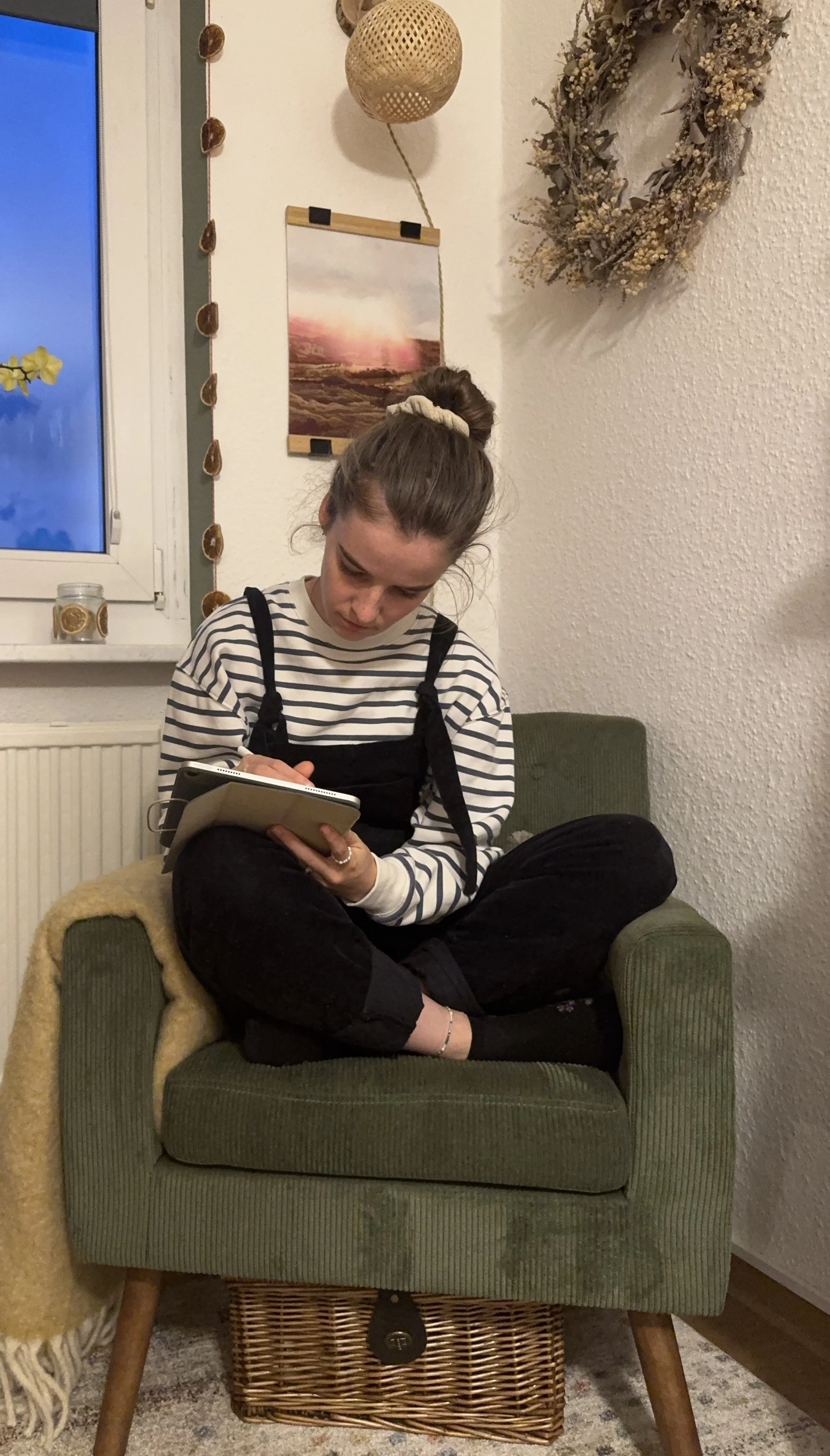 Young woman sitting cross-legged on a green armchair, writing in a notebook. She is in a cozy room with potted plants, wall art, and decorative items. A blanket drapes over the chair's arm. The room has a rustic and warm ambiance.