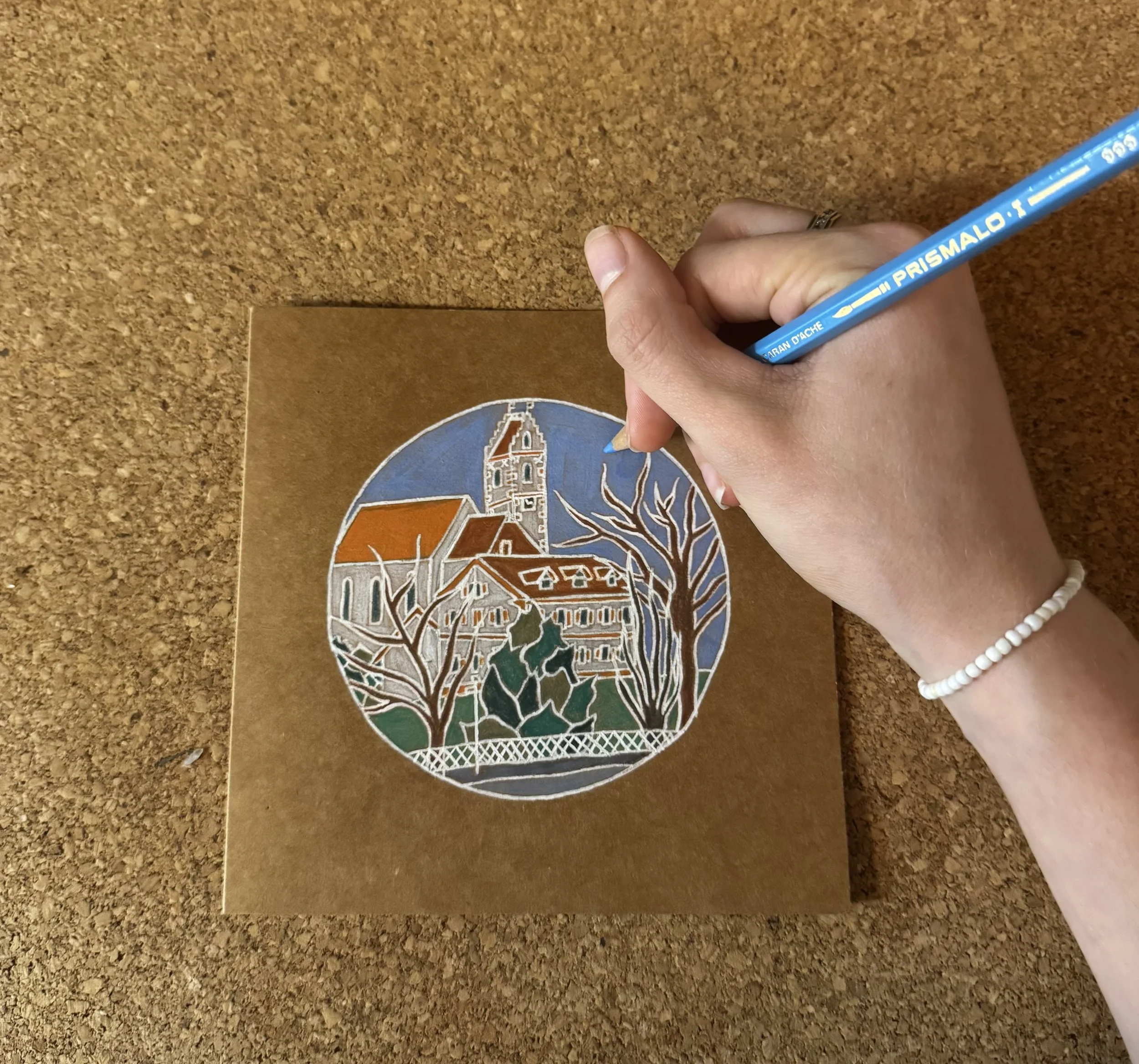 A hand holding a blue Prismalo colored pencil writing on brown paper with a colored circular illustration of a building and landscape.