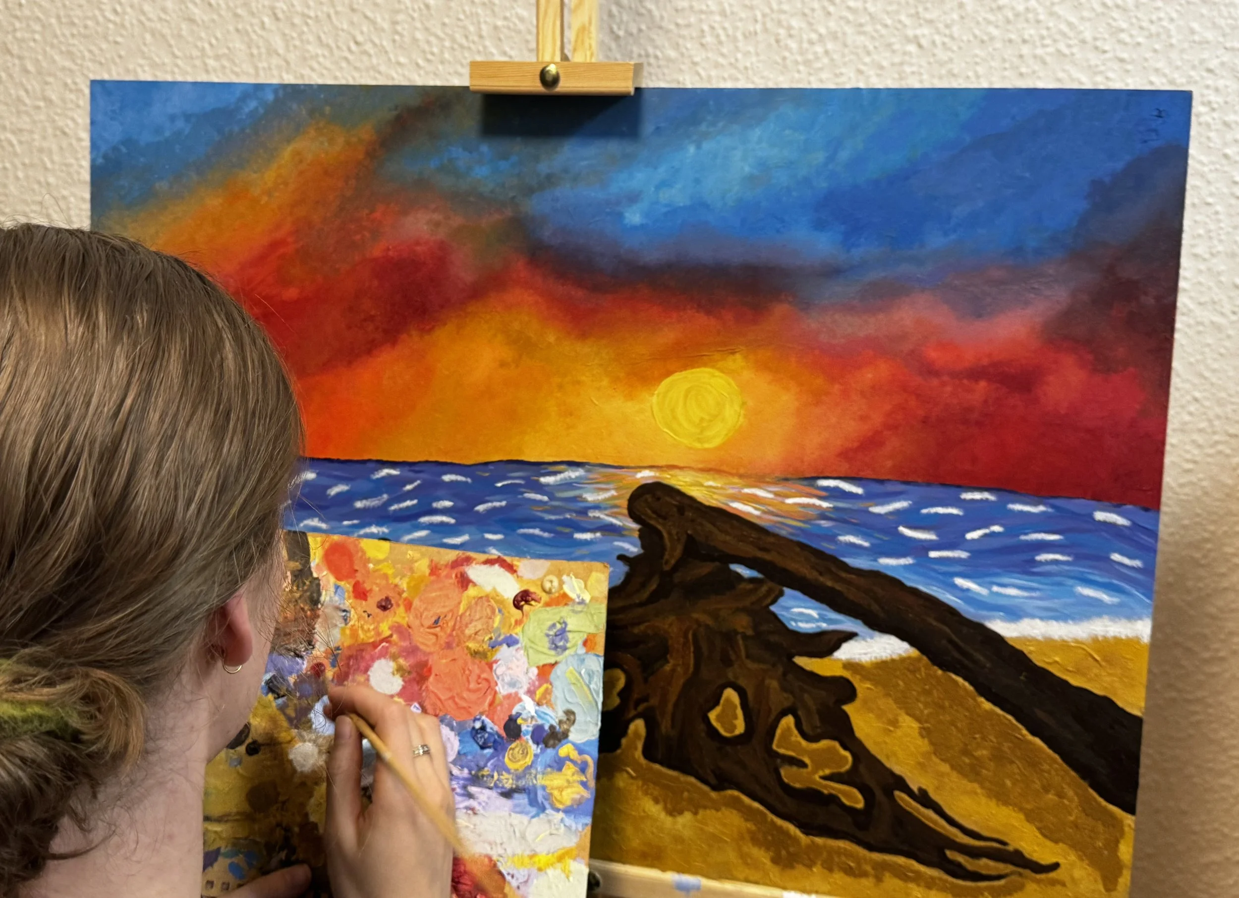 Artist painting a vibrant sunset scene with a tree silhouette on a colorful landscape canvas.