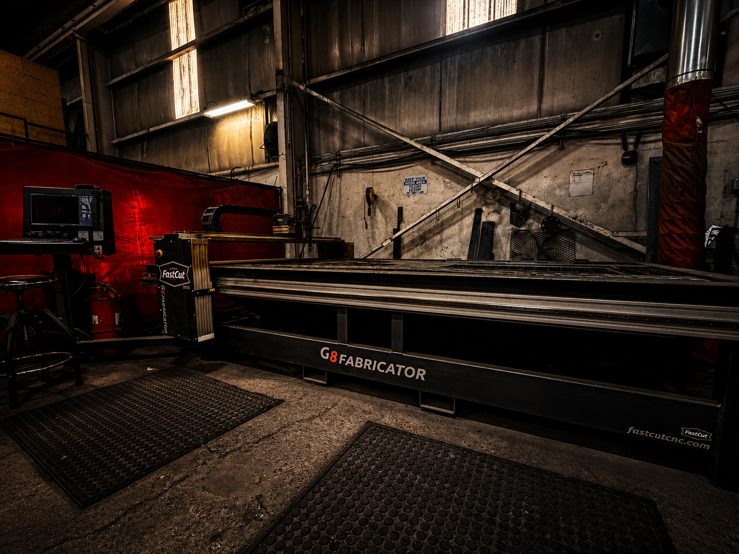 An industrial laser cutting machine in a workshop with a computer monitor on a stand and rubber mats on the floor.
