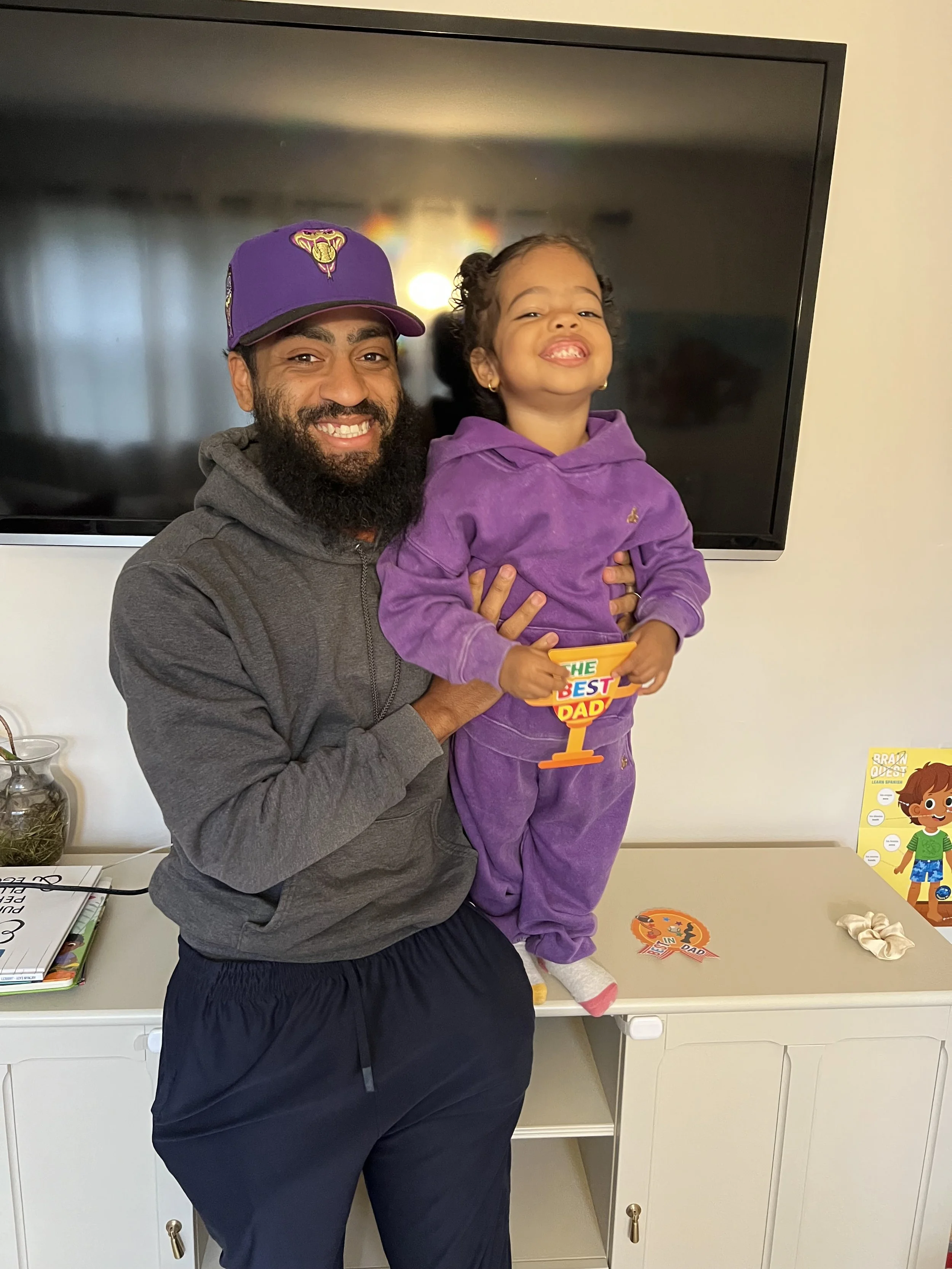 A man and a young girl smiling and posing together indoors. The girl is holding a colorful sign that says "The Best Dad". Behind them, a TV is mounted on the wall, and there are various items on a white cabinet, including books and decorations.