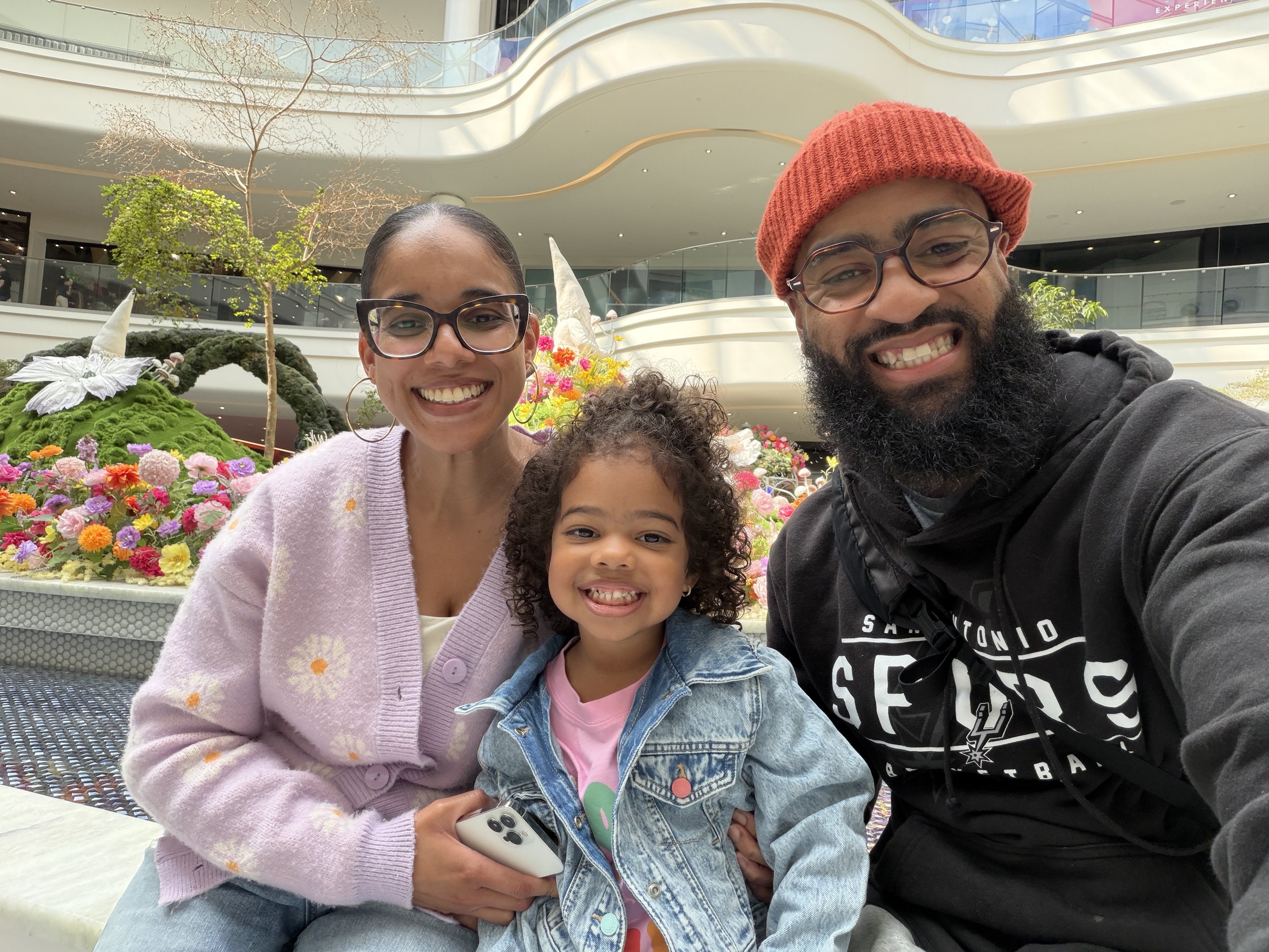 A smiling woman, a young girl, and a man taking a selfie together in an indoor shopping mall with floral displays and modern architecture.