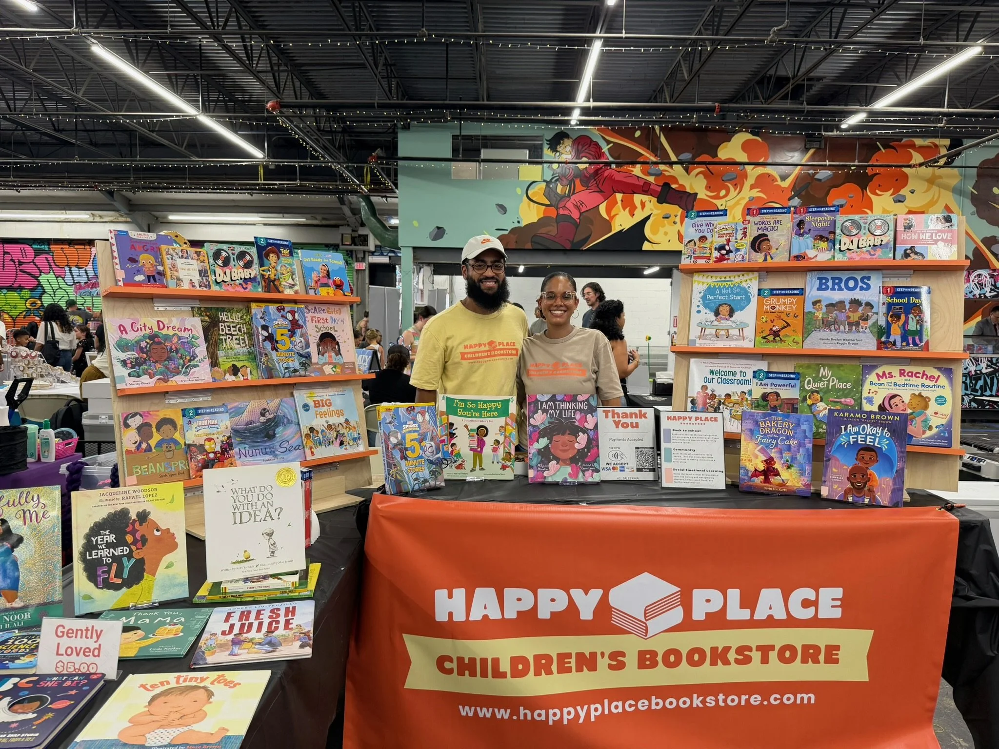 Children's books displayed on tables at a bookstore event with a colorful graffiti mural of a person in combat gear on the wall behind. Two smiling people stand behind the table in front of the books, under a string of lights.