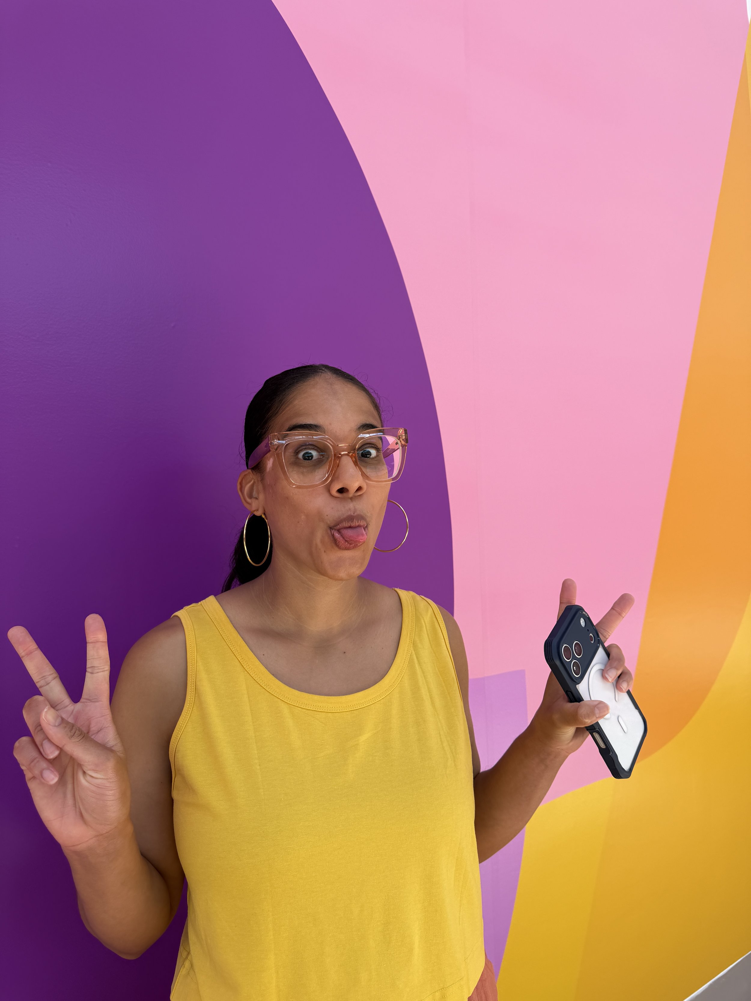 A woman in a yellow tank top and pink glasses making a peace sign with one hand and holding a smartphone with a game controller grip in the other hand, against a colorful abstract background of purple, pink, and yellow.
