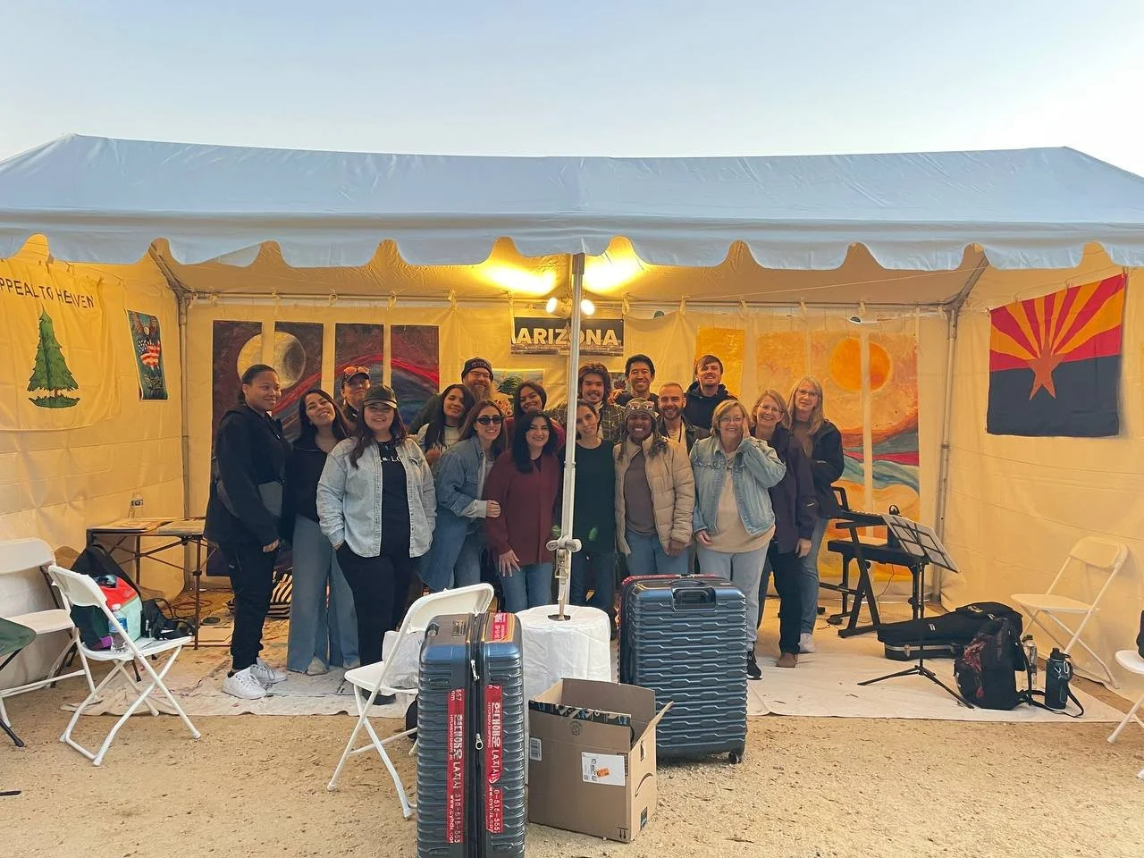 Group of people standing together inside a large tent decorated with artwork and flags, with musical equipment and luggage in the foreground.