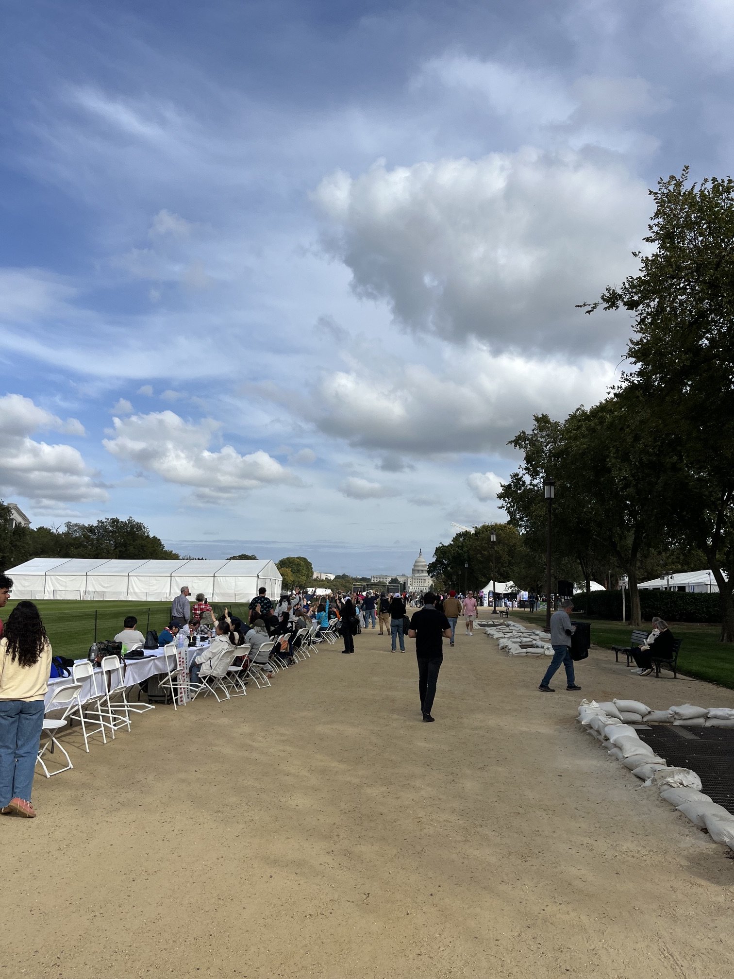 Outdoor event near the U.S. Capitol with tables, chairs, and people, under a partly cloudy sky, with tents and sandbags along the path.