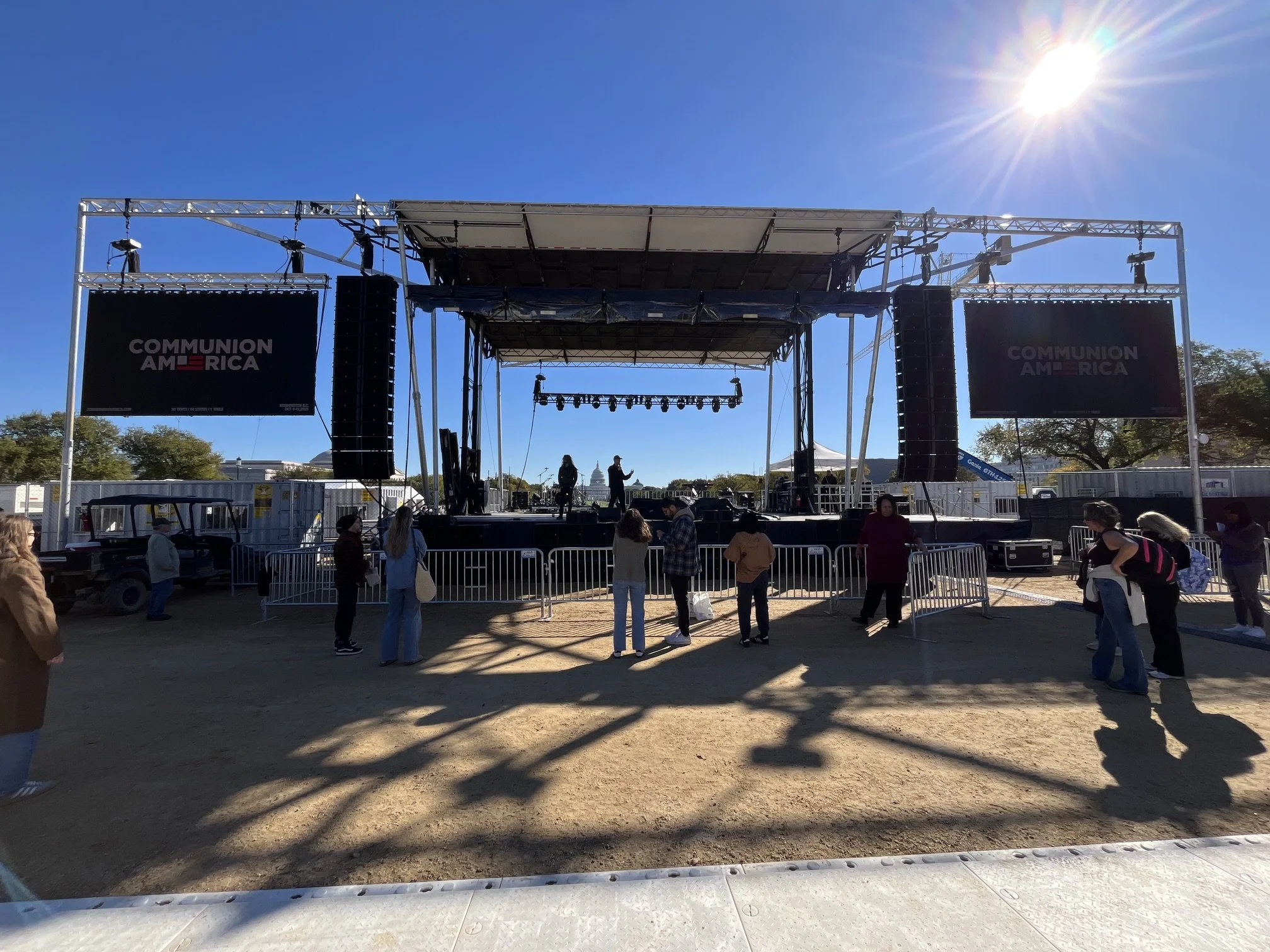 Empty outdoor stage with large screens displaying the words 'Communion America', set against a clear blue sky with the sun shining brightly.