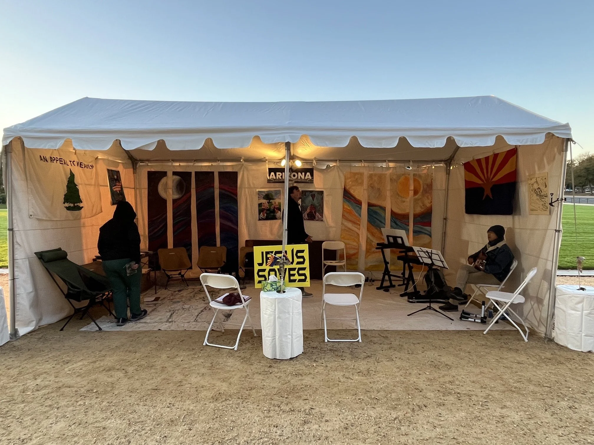 A white event tent set up outdoors with people inside, some playing guitar, and artwork displayed on the walls, including a sign that says 'Jesus Saves' and a flag with a tree, a sun, and a mountain.