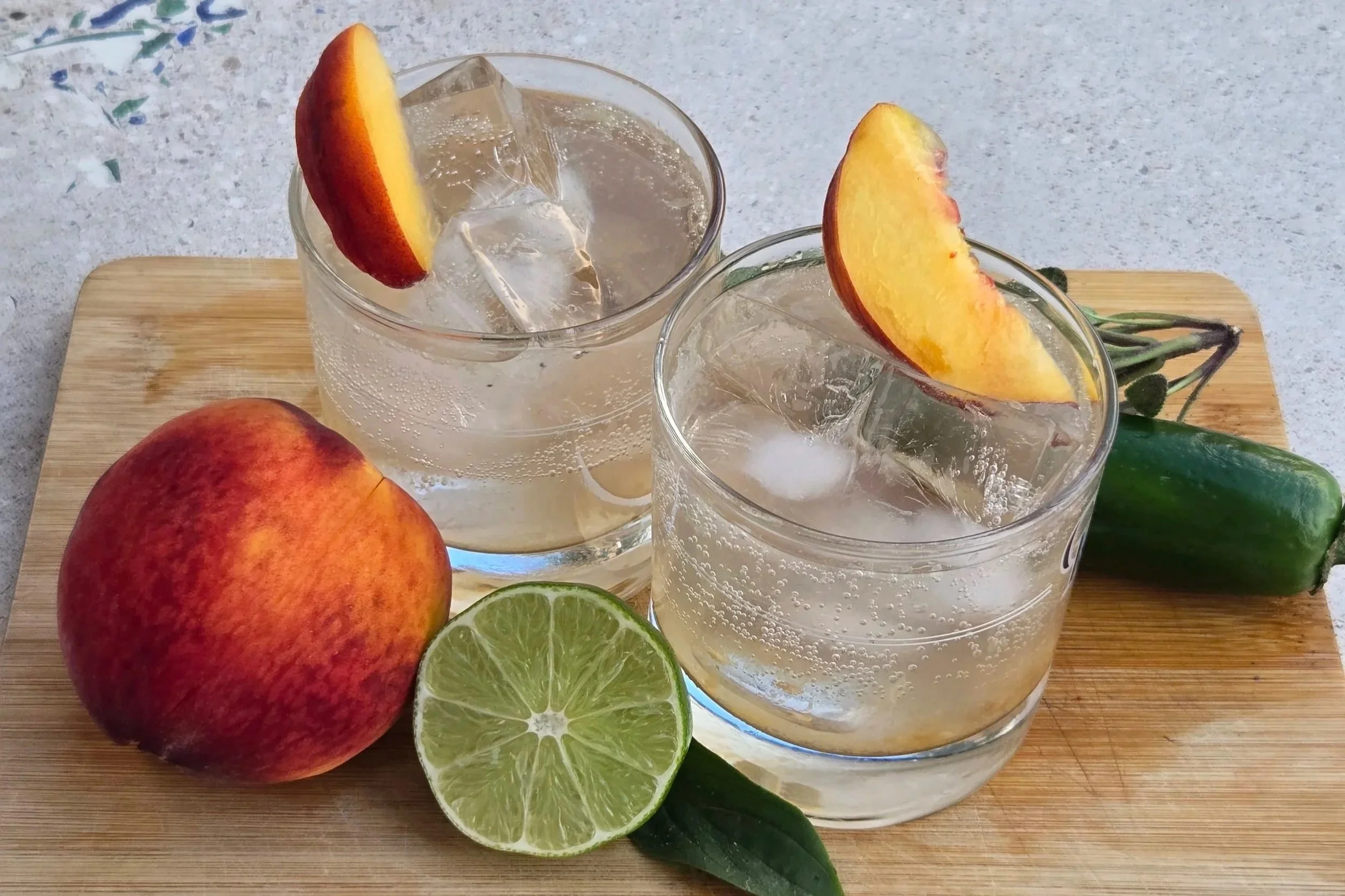 The Palisade Picoso. Two glasses of clear beverage with ice and peach slices, a whole peach, a halved lime, a cucumber, and some green leaves on a wooden cutting board.