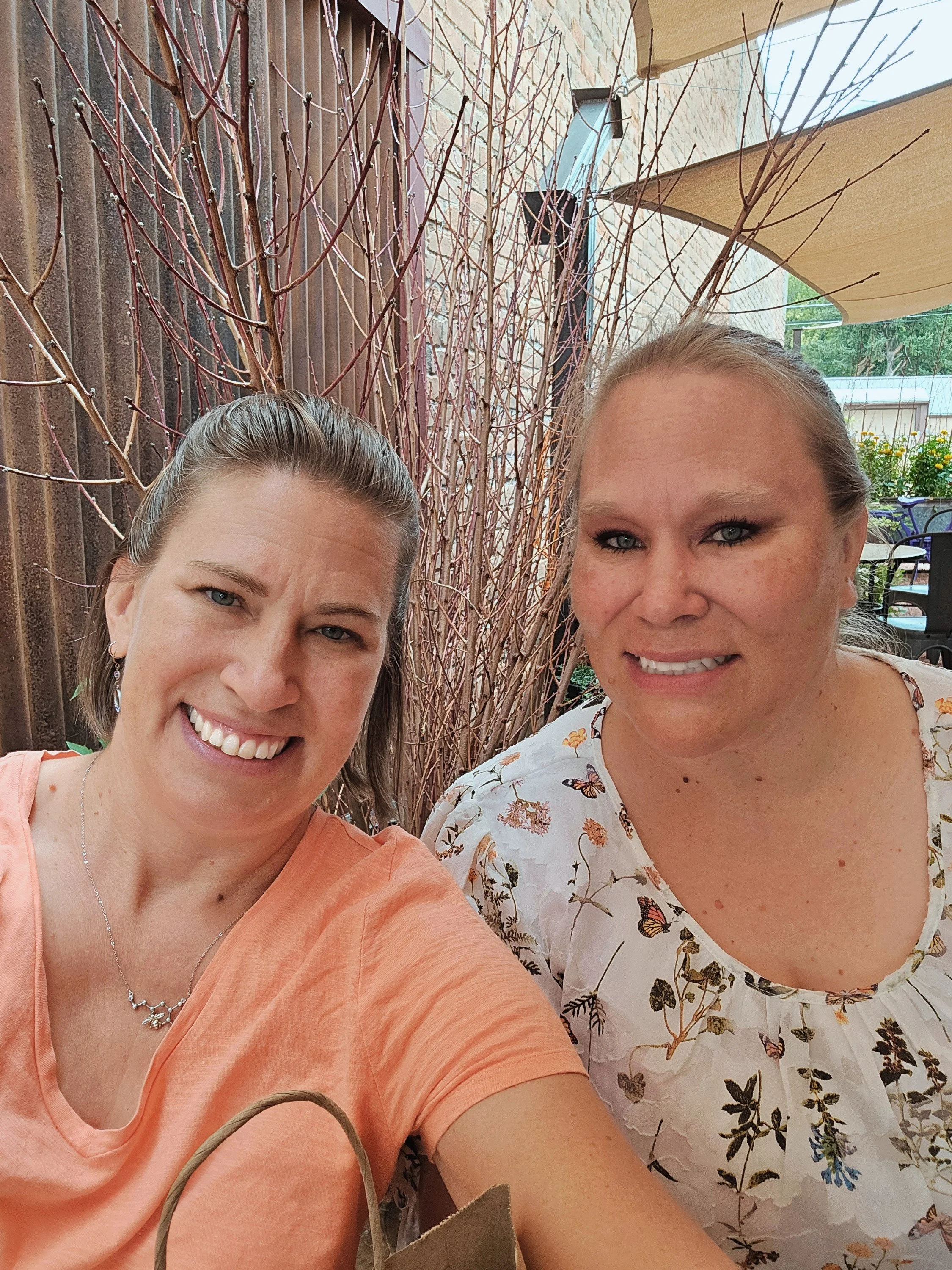 Two sisters, smiling in an outdoor setting at Sage Creations, in Palisade, Colorado. Background has a brick wall, wooden fence, greenery, and patio umbrellas in the background.