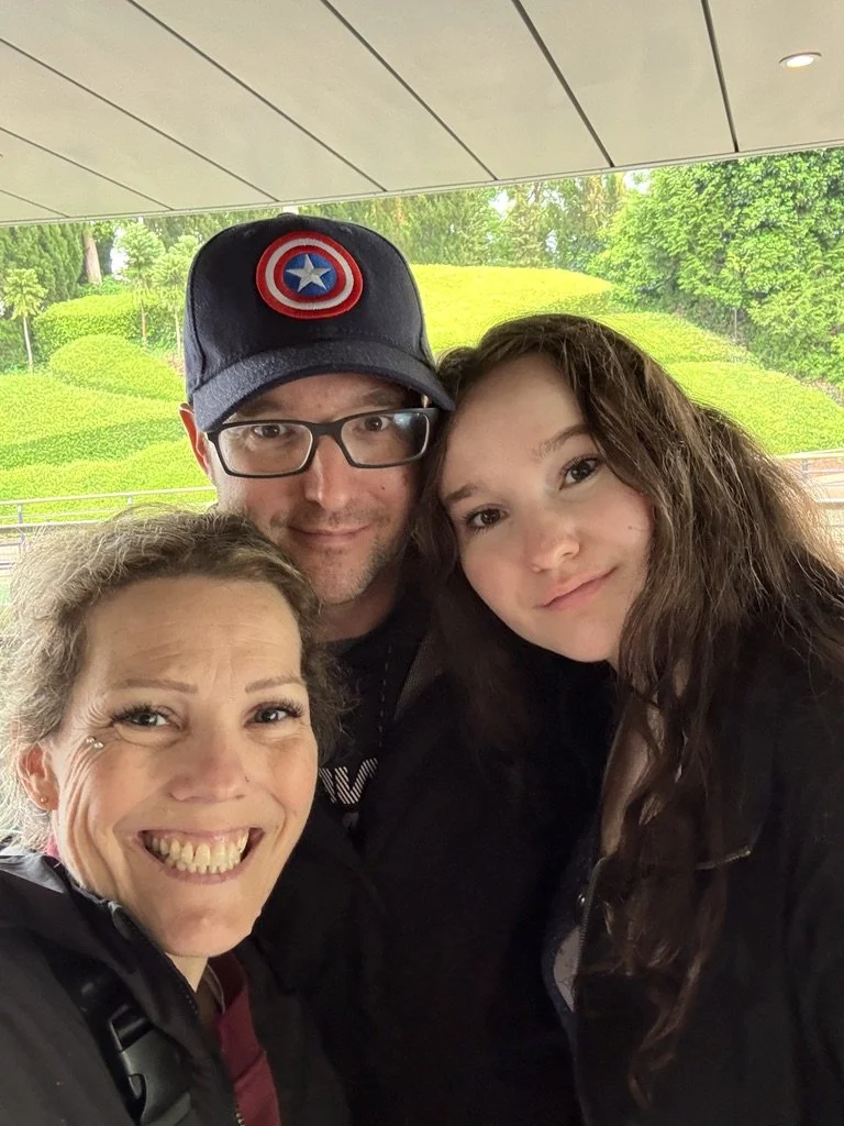 A group of three people smiling for a selfie outdoors under a roof, with green, hilly landscape and trees in the background.
