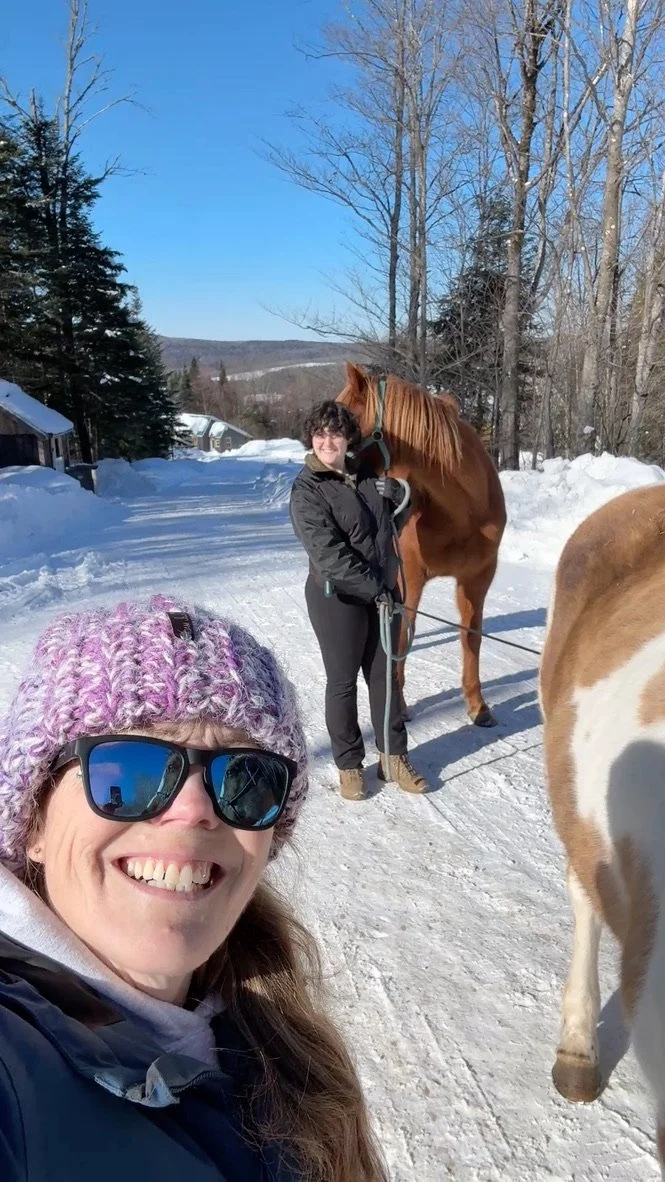 Two women in winter clothing smiling on a snowy trail, one taking a selfie and the other holding a horse, with trees and a clear blue sky in the background.