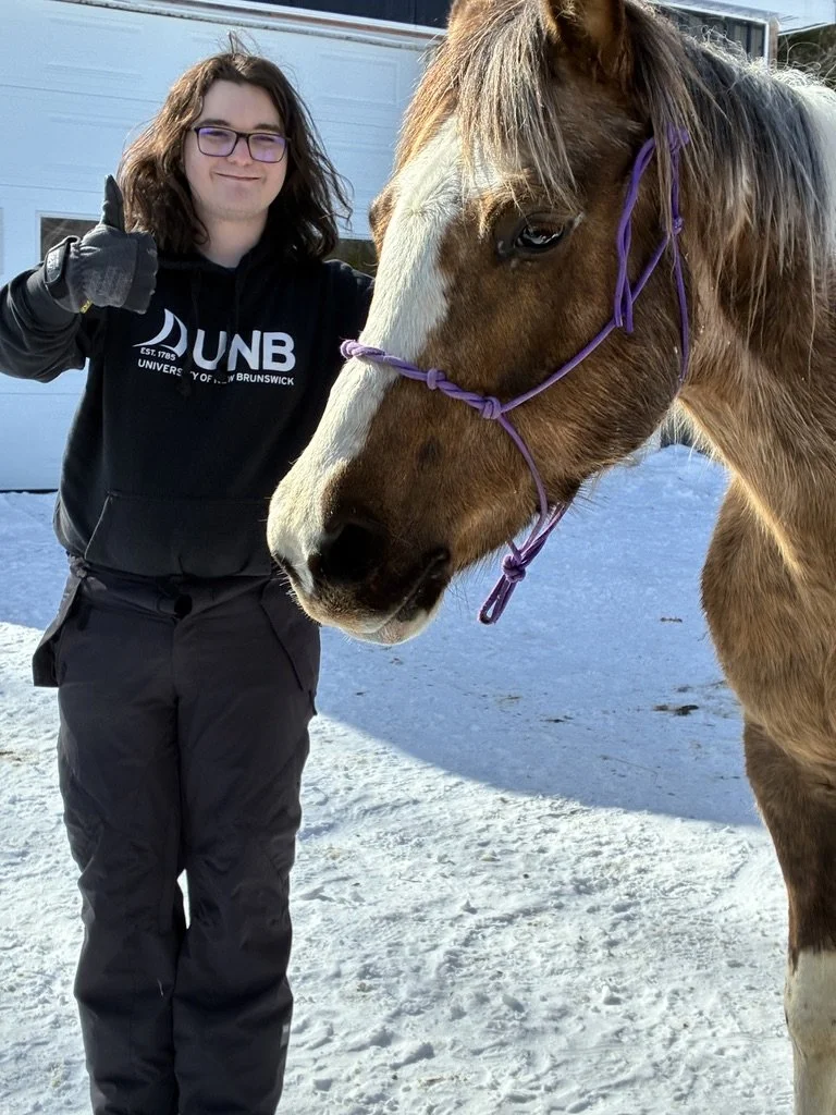 A person standing outdoors in winter clothes, giving a thumbs-up, next to a horse with a purple halter.