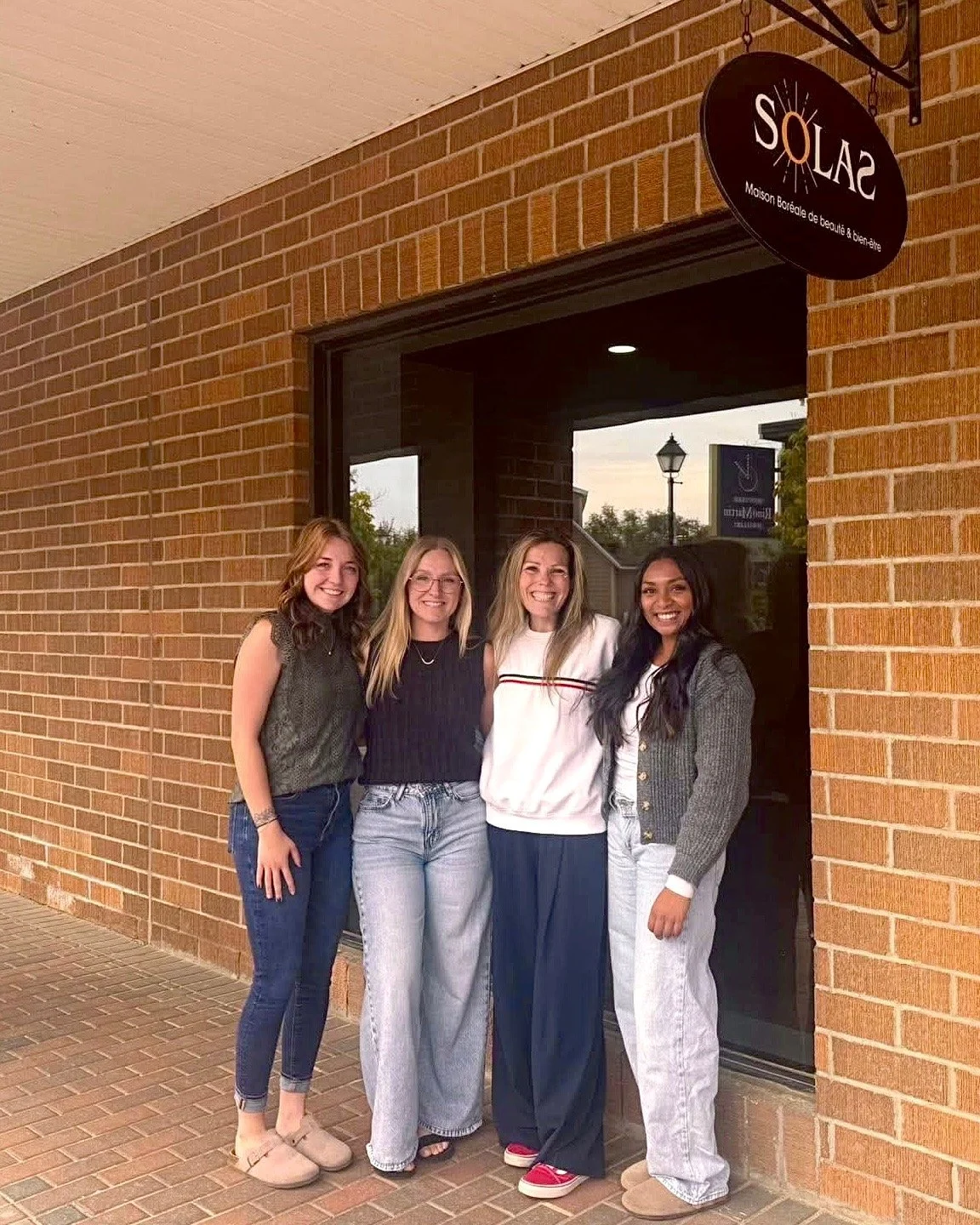 Four women standing outside a brick building, smiling at the camera, in front of a sign that reads "SOLAS" with additional text below.