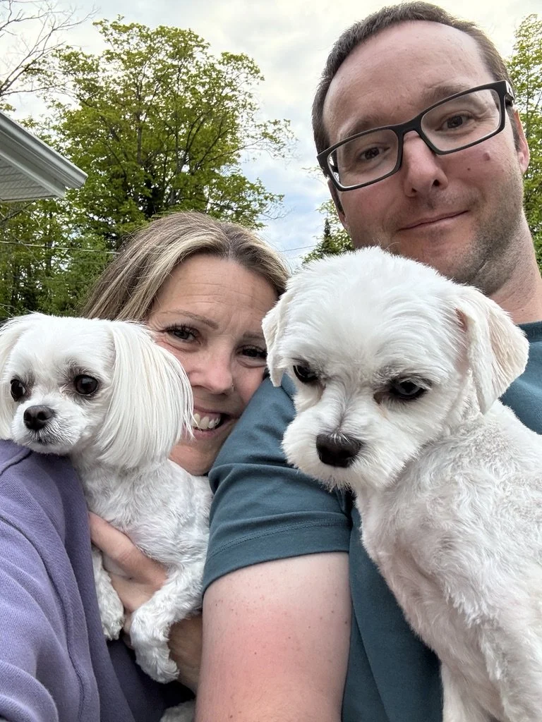 A man and woman taking a selfie outdoors with two small white dogs, surrounded by green trees and a partly cloudy sky.
