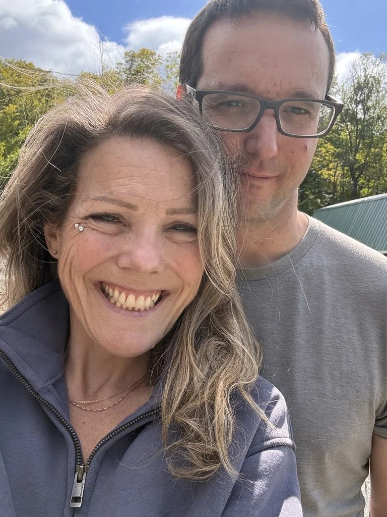 A woman with wavy blonde hair smiling and a man with short brown hair and glasses standing outdoors on a sunny day.