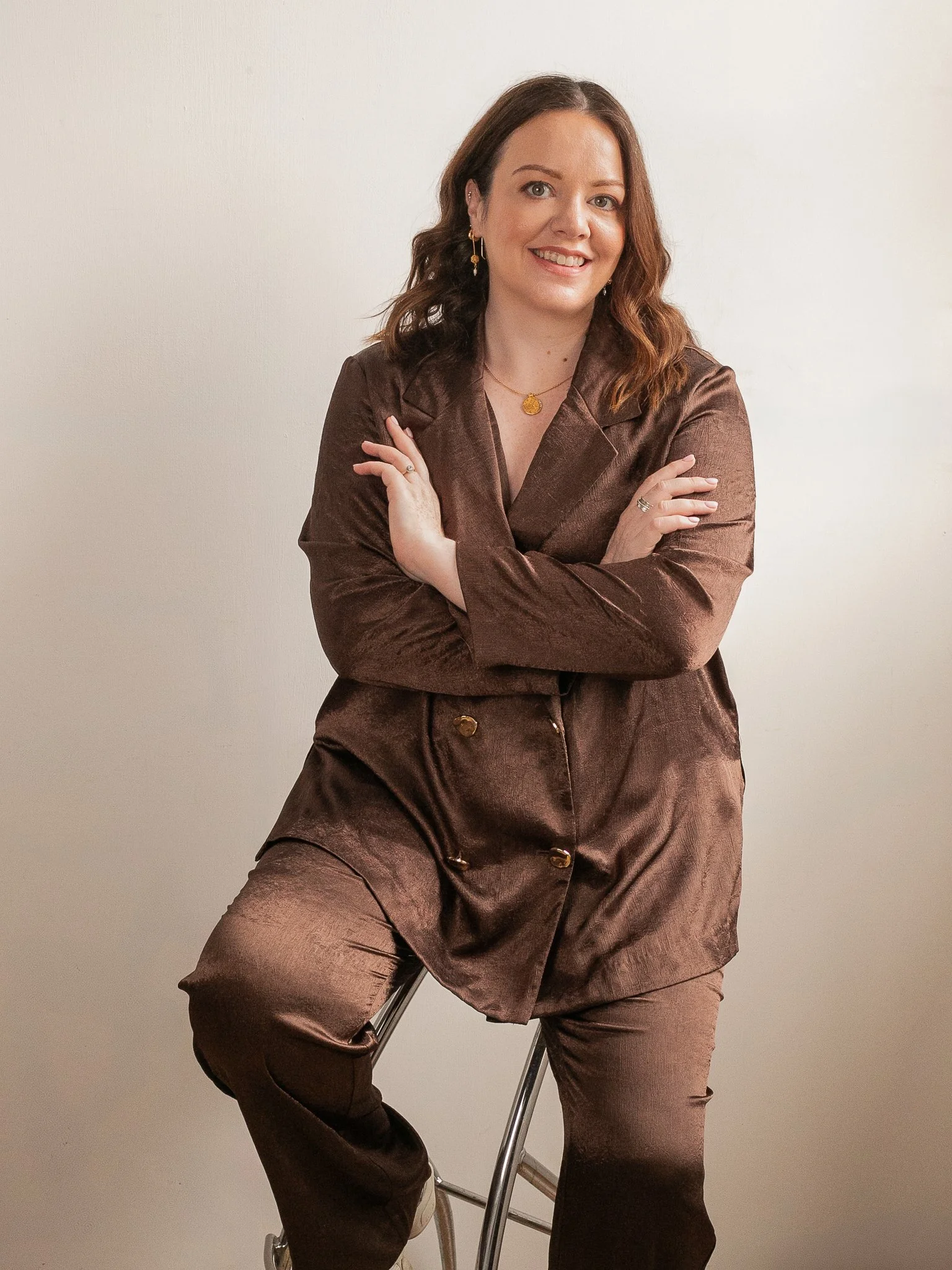 Portrait of an Irish ADHD coach wearing a warm brown satin suit, seated on a stool and smiling against a neutral background.