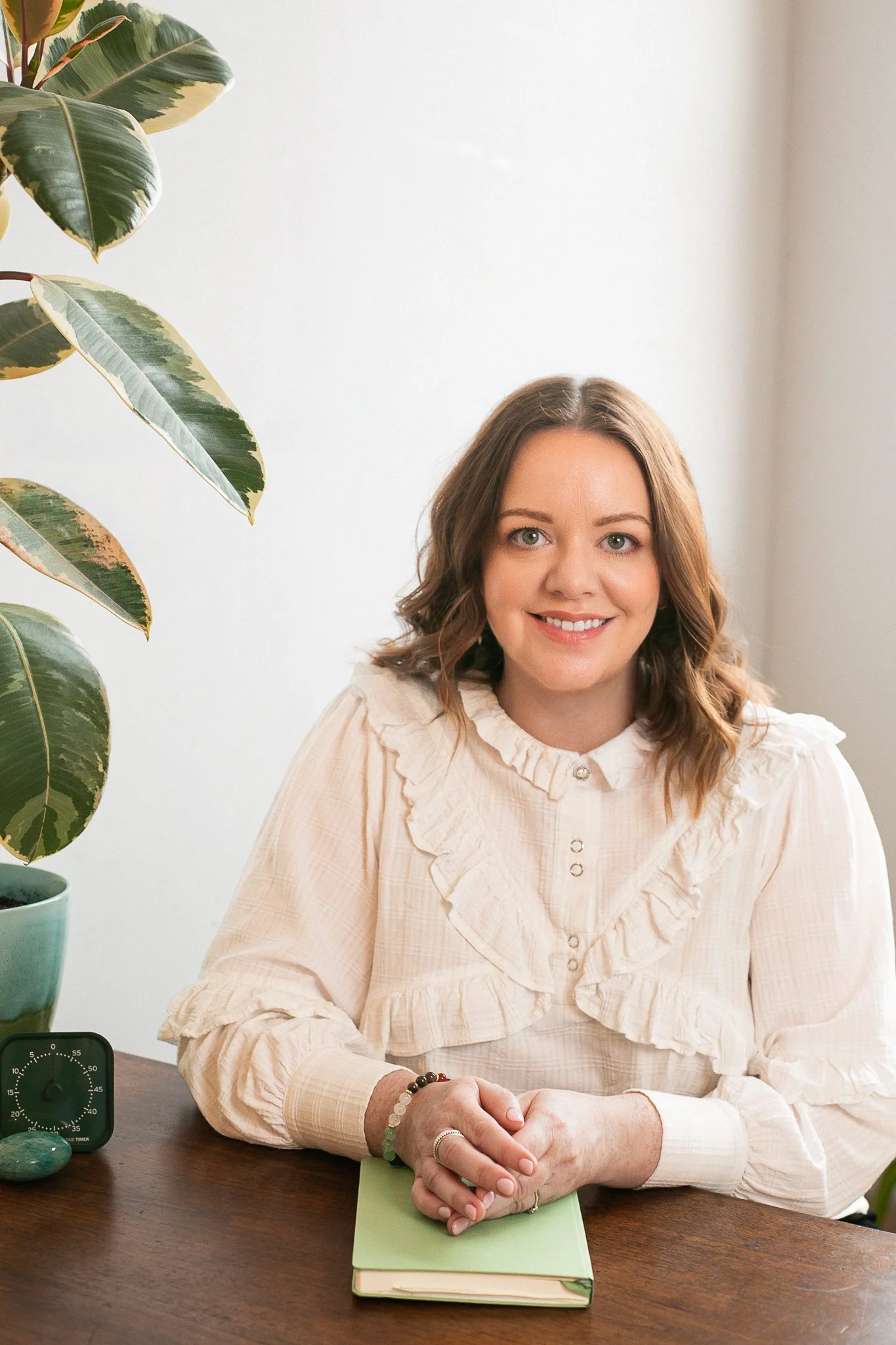 An Irish female ADHD coach sitting at a desk with her hands folded, wearing a white blouse with a plant, visual timer and notebook on the desk.