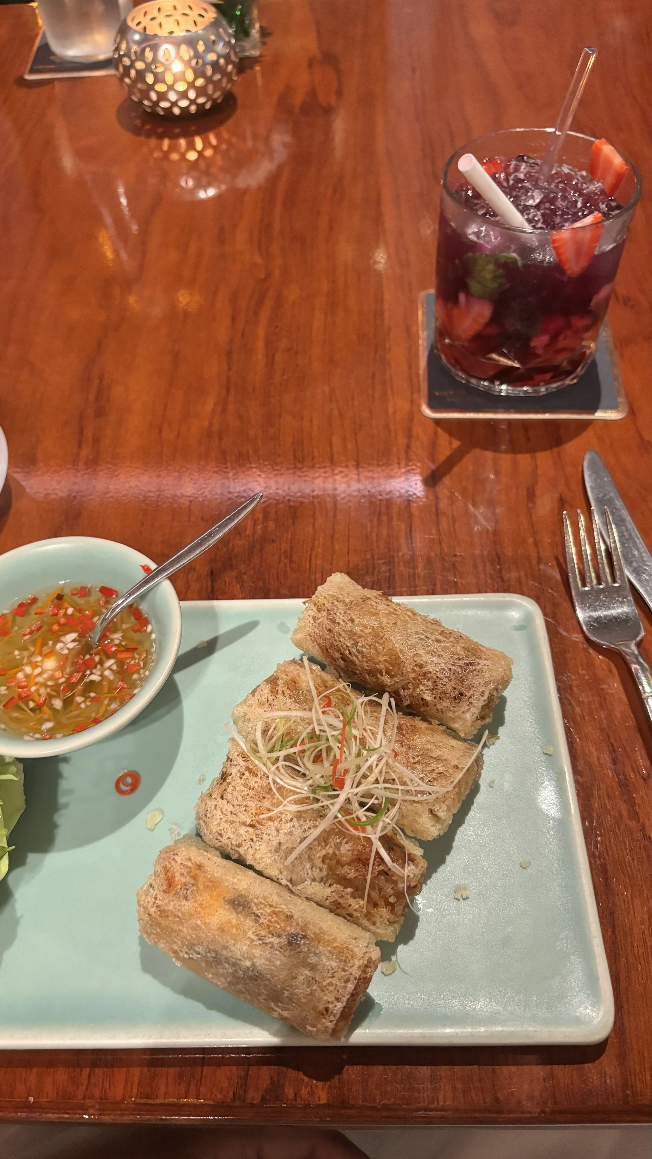 A plate of spring rolls with a small bowl of dipping sauce, a chili garnished topping, on a mint green plate. A drink with strawberries, mint, and ice, with straws, on a coaster. The table is wooden, with a small candle holder in the background.