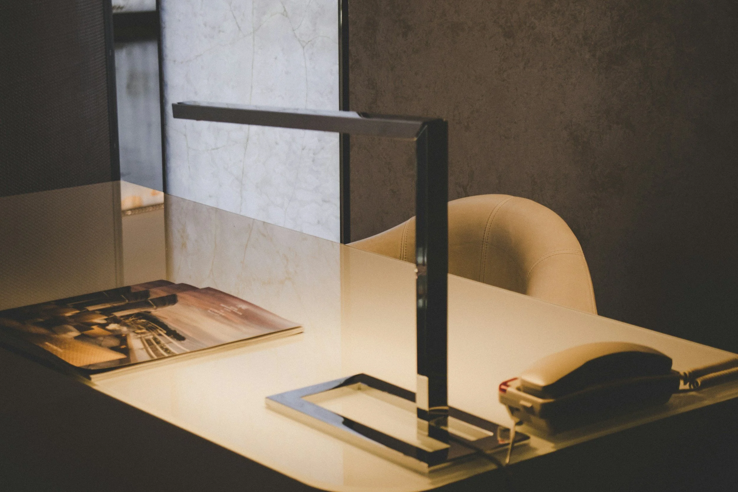 A workspace with a white table holding a recent magazine and some papers, separated by a black metal divider, with a beige upholstered chair and a marble-top surface in the background.