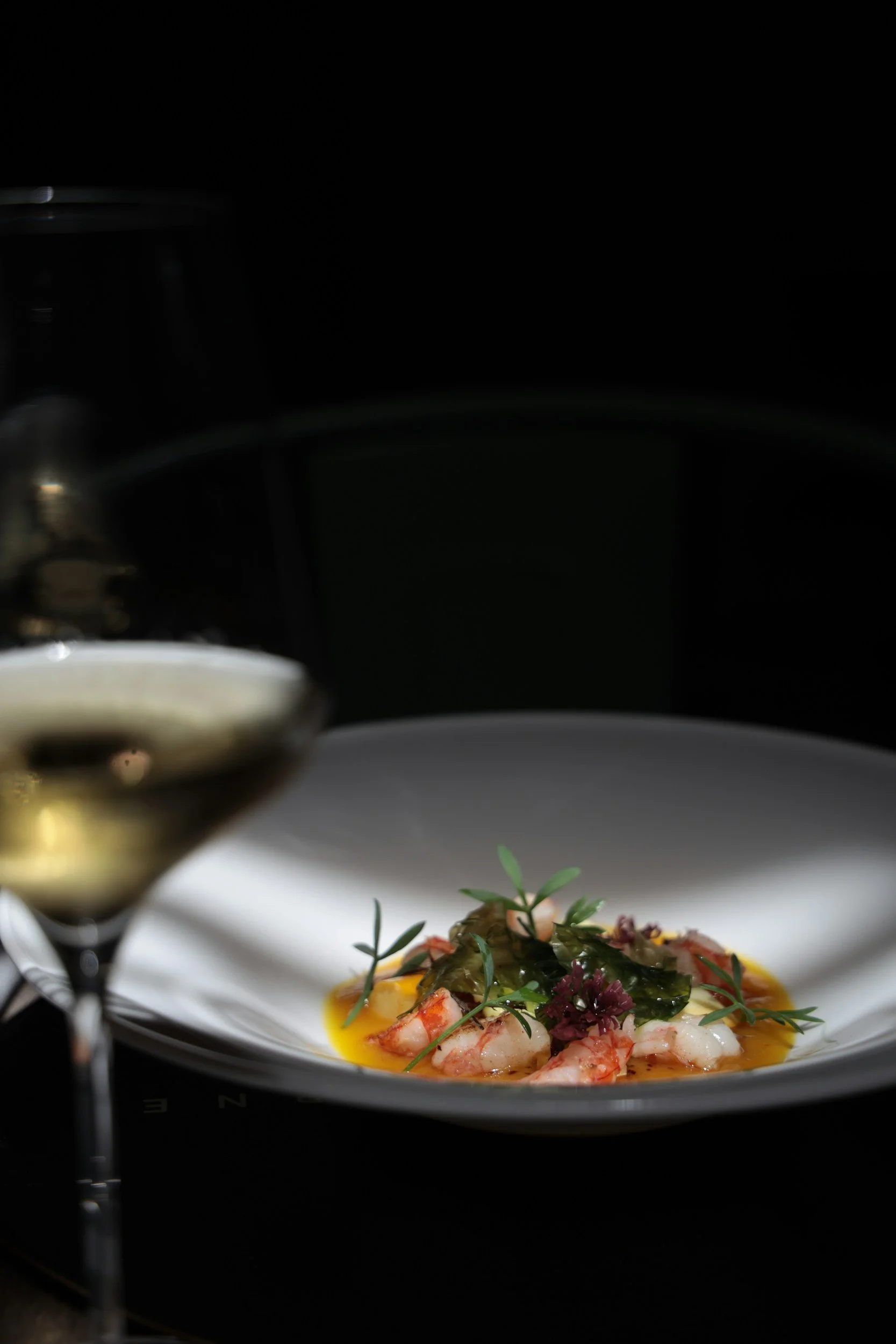 A plate of gourmet food featuring shrimp, garnished with herbs and vegetables, with a mustard-colored sauce and a drizzle of oil, in a fine dining setting.