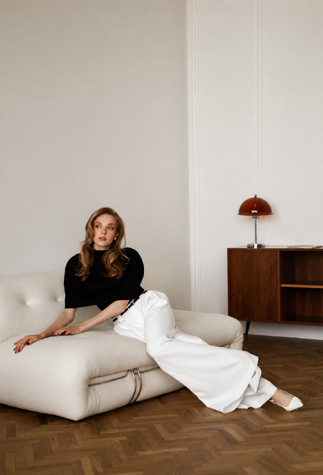 A woman with blonde hair, wearing a black top and white pants, sitting on a cream-colored sofa in a minimalist room with wooden flooring and a wooden sideboard with a red desk lamp.
