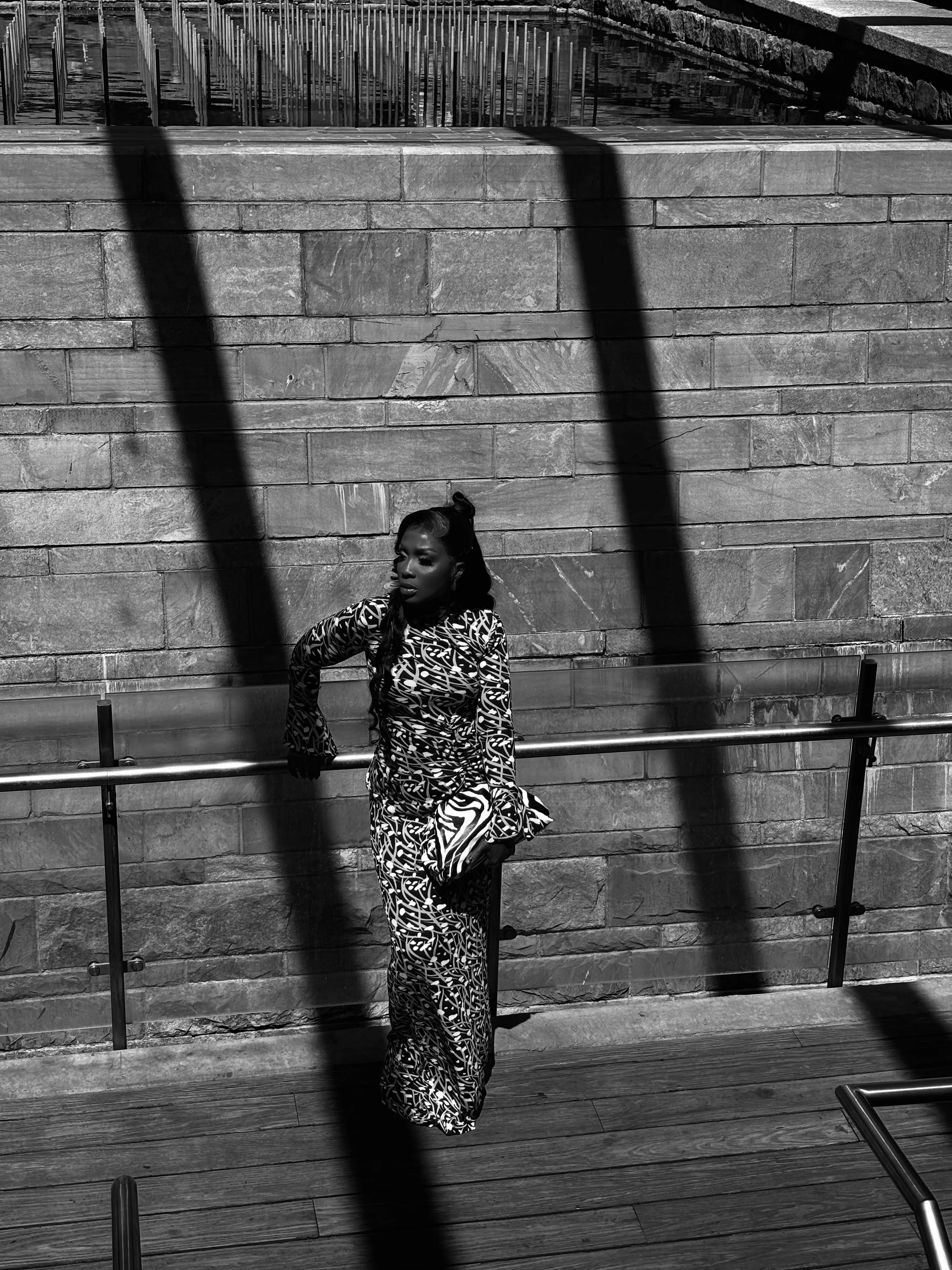 A woman in a patterned dress stands against a brick wall with shadows cast across her and the scene. She leans on a metal railing, with wooden flooring at her feet.