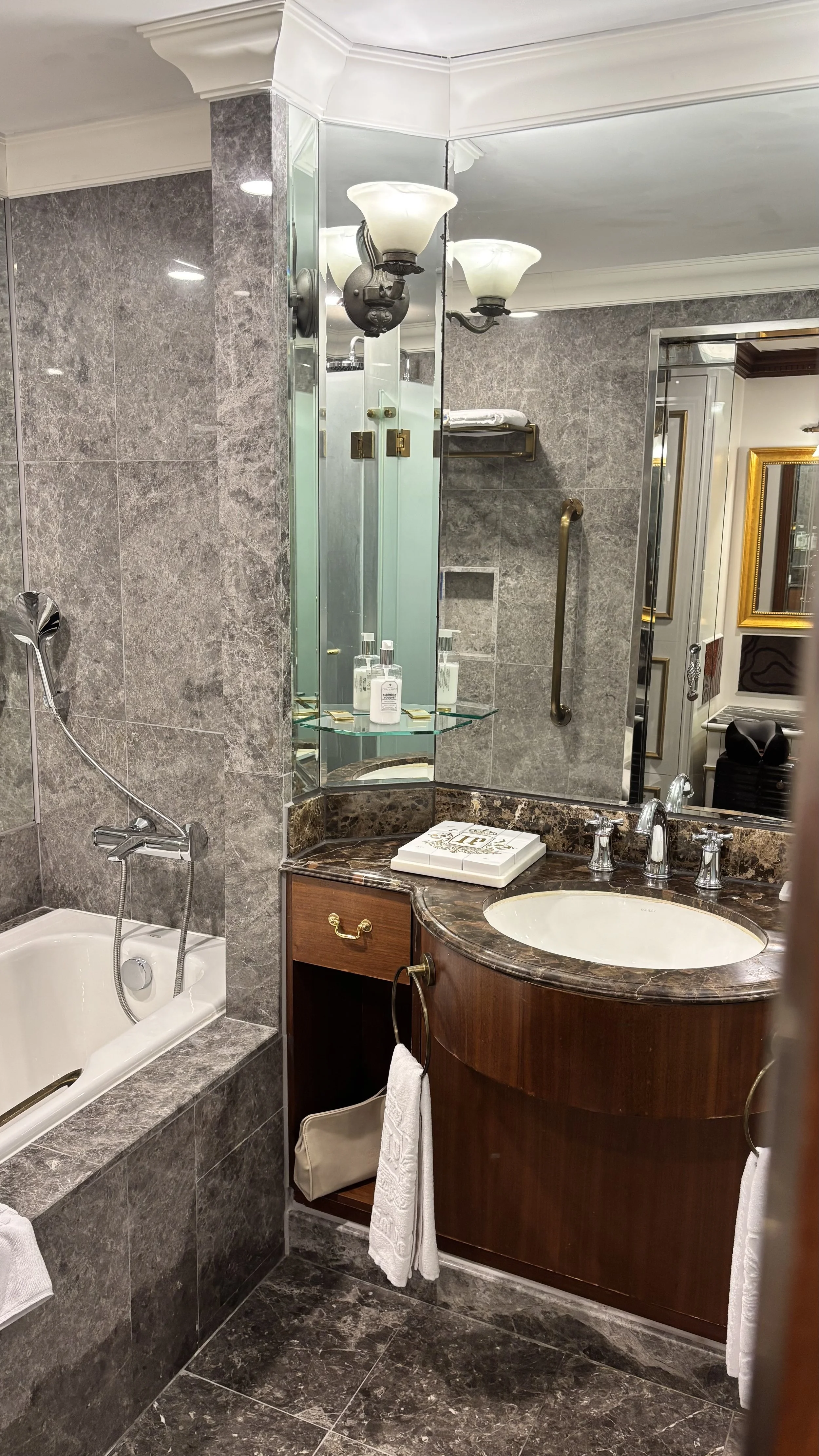 A luxurious bathroom with a bathtub, a sink with a marble countertop, a mirror, and decorative lighting. The walls and floors are covered in gray marble tiles, and there are glass shelves with toiletries. The bathroom has elegant fixtures and gold accents.