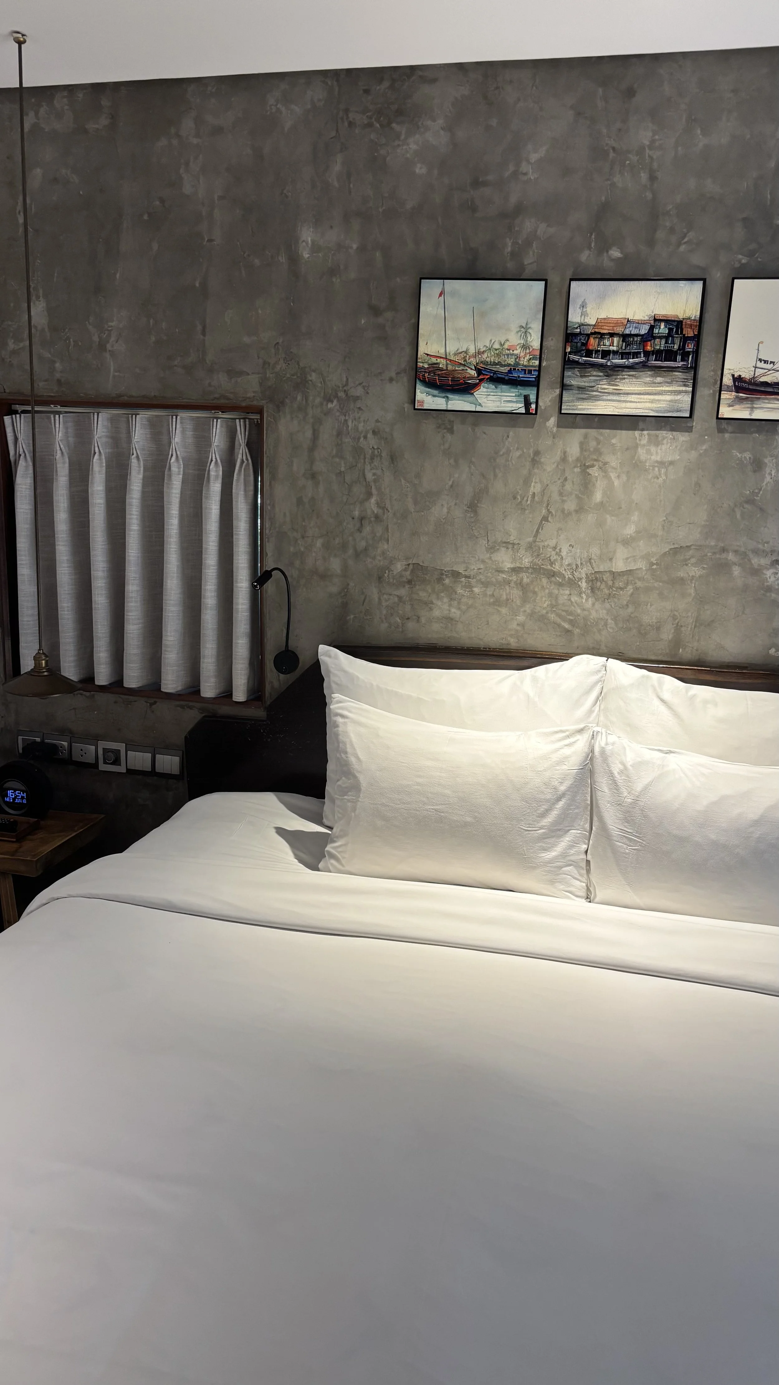 A bedroom with a concrete wall, three framed paintings of boats and water, a window with striped curtains, a black lamp, a bedside table with a clock, and a bed with white pillows and sheets.