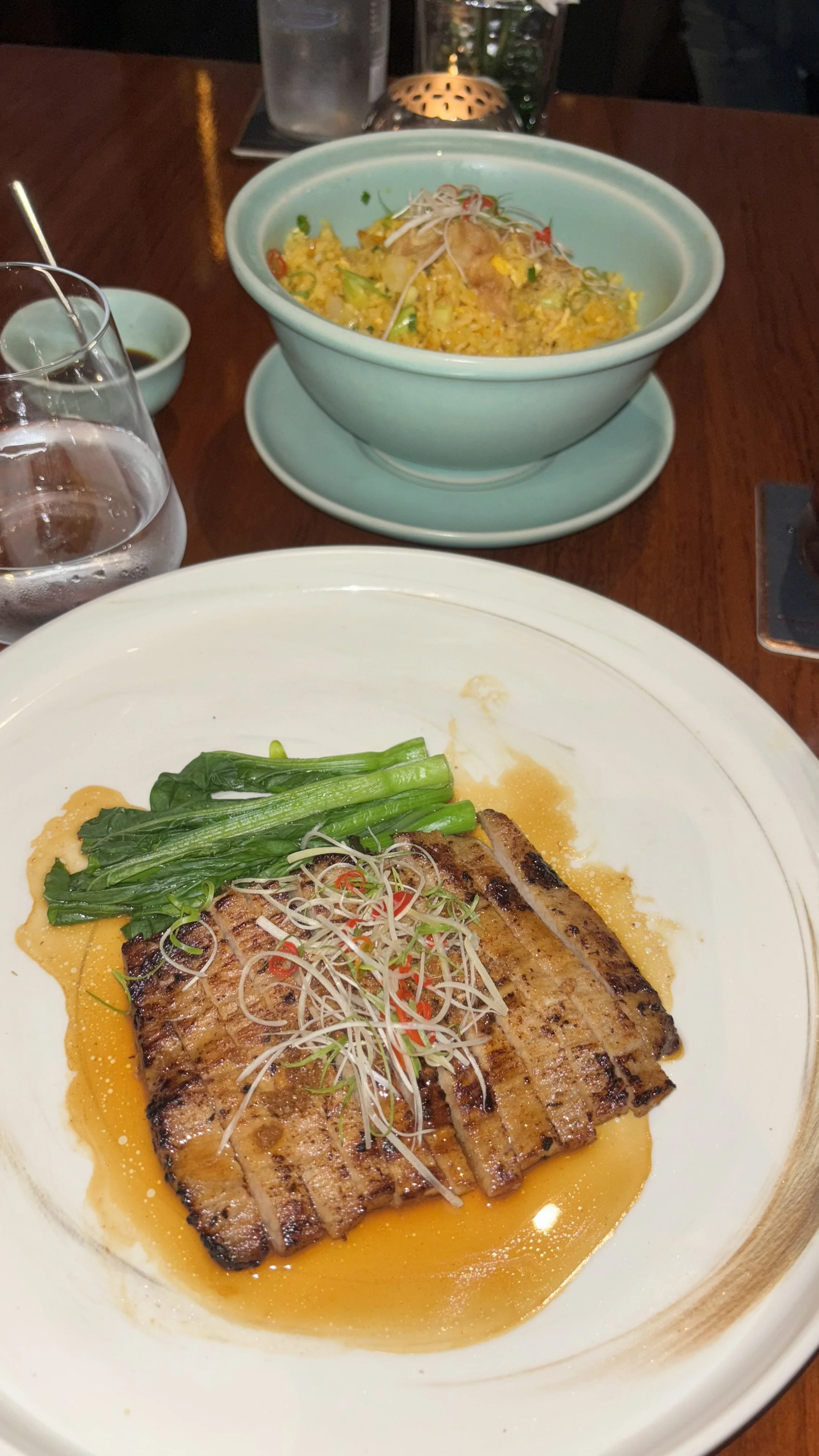 A plate with grilled pork belly, green vegetables, and sprouts, topped with sliced red chili peppers. In the background, there is a bowl of fried rice with vegetables and garnishes.
