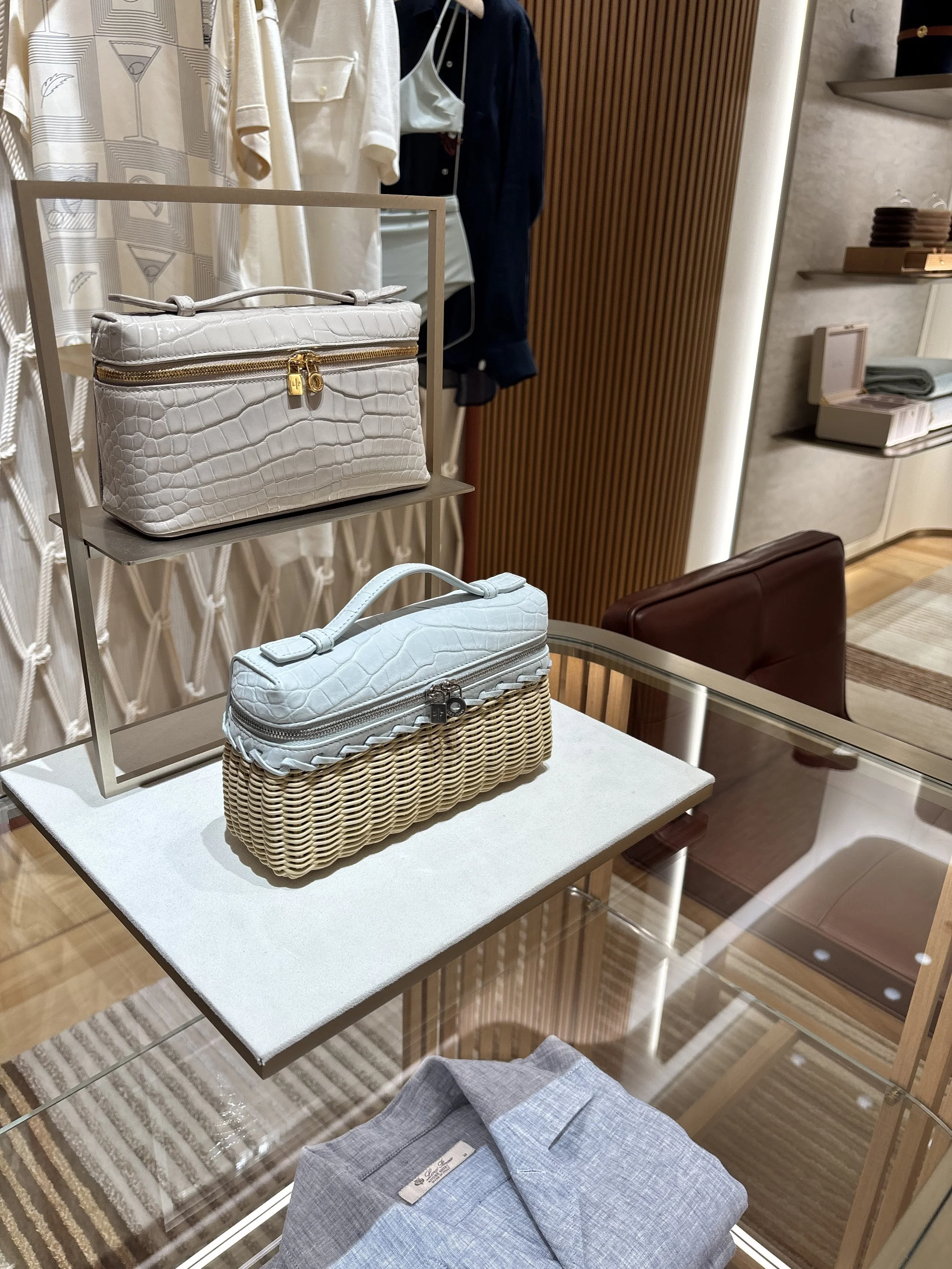Display of various handbags, including a white crocodile-textured clutch with a gold zipper and lock, and a light blue textured clutch, placed on a white table inside a retail store.