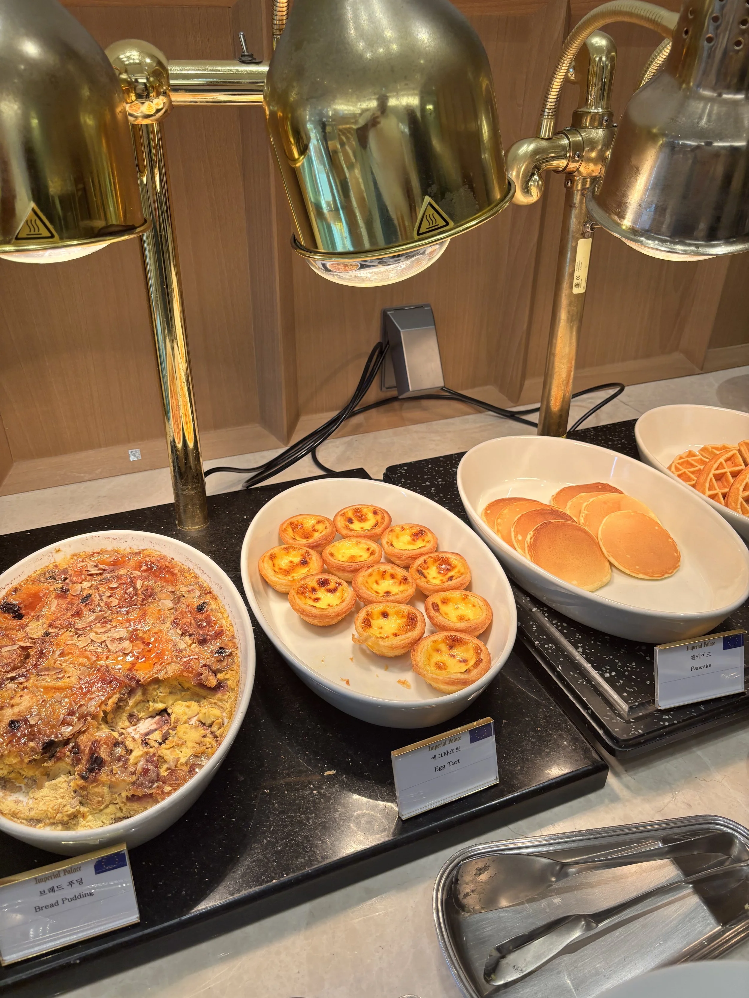 Buffet breakfast setup with various dishes including bread pudding, egg tart, pancakes, and waffles with chafing dishes and small signs labeling each dish.