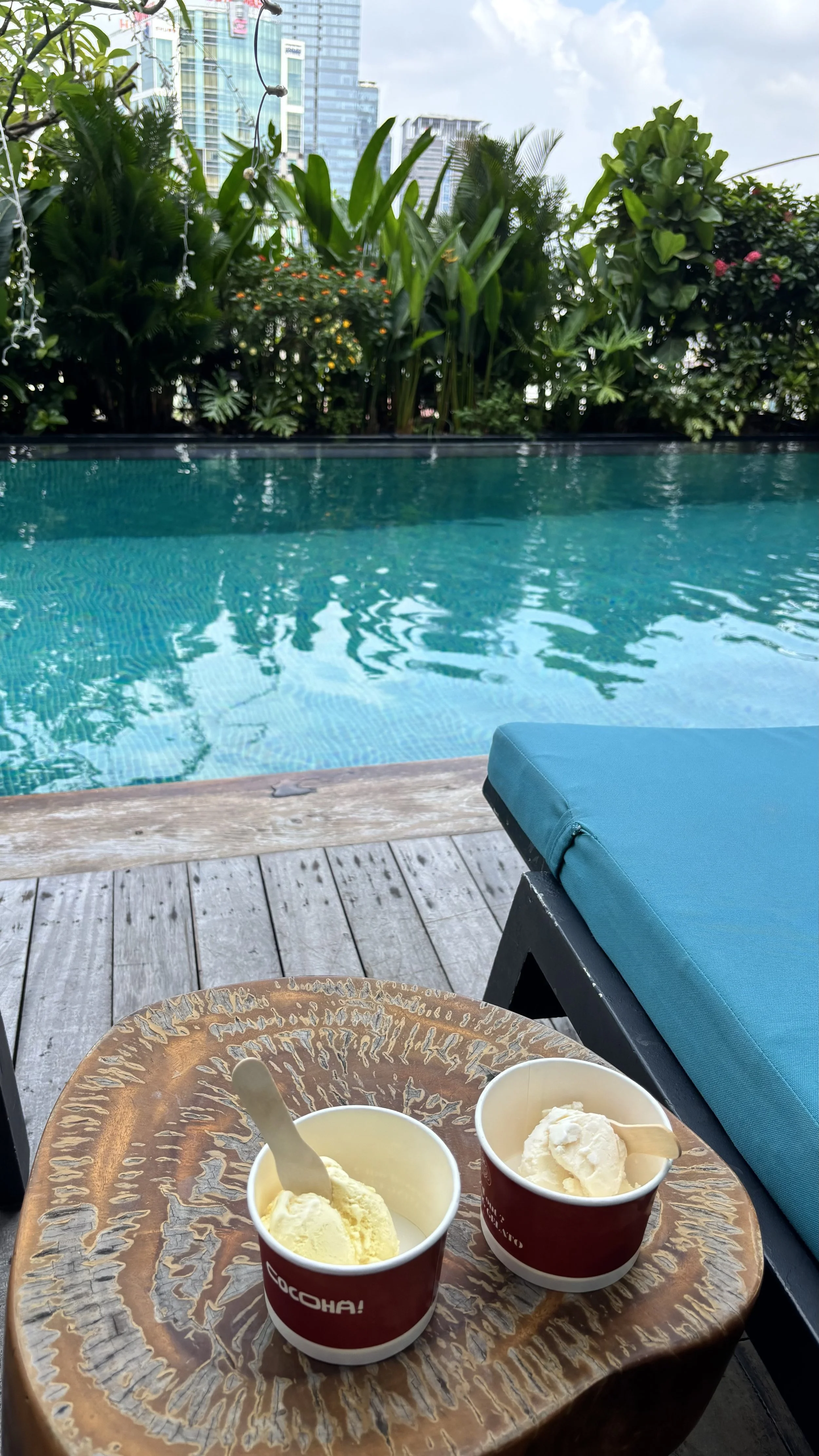 Two cups of vanilla ice cream with small wooden spoons sit on a decorative wooden table next to a swimming pool, with lush greenery and tall buildings in the background on a cloudy day.