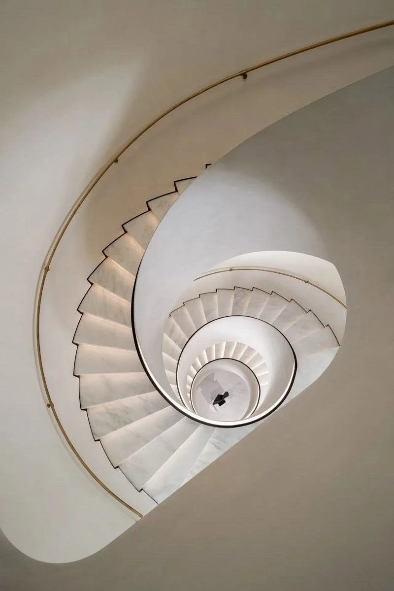 A top-down view of a spiral staircase with white marble steps and a gold handrail, descending into a circular opening at the bottom.