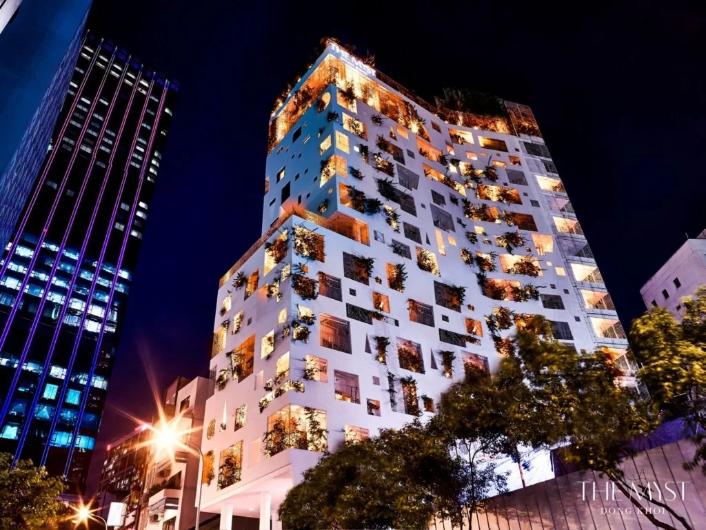 Nighttime cityscape featuring a modern white building with irregularly shaped balconies and greenery, adjacent to a tall dark building with lit windows and vertical neon lights.