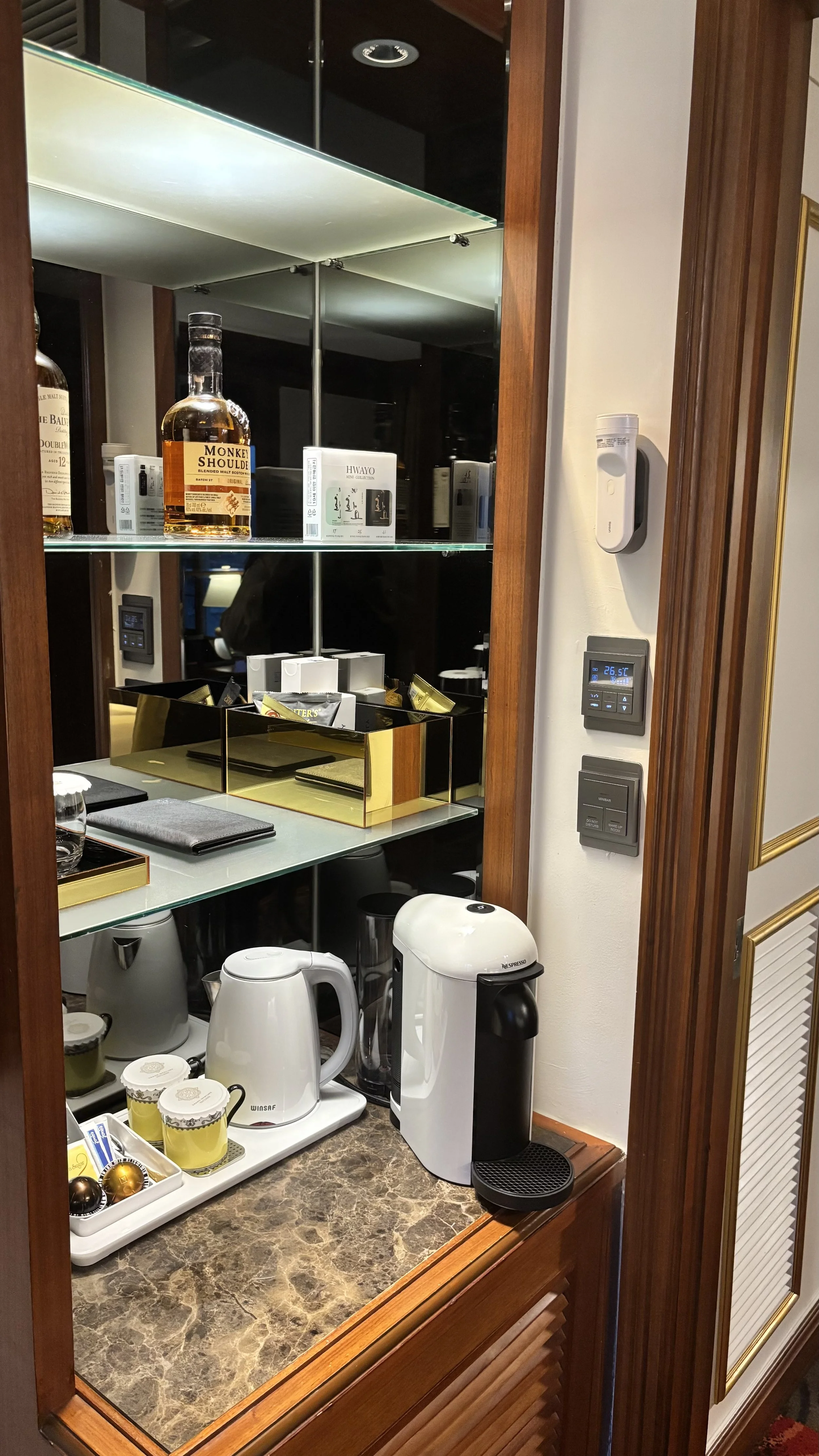 A coffee station with various appliances and beverages, including a Nespresso machine, electric kettles, and cups, with a marble countertop and mirrored shelves behind it.