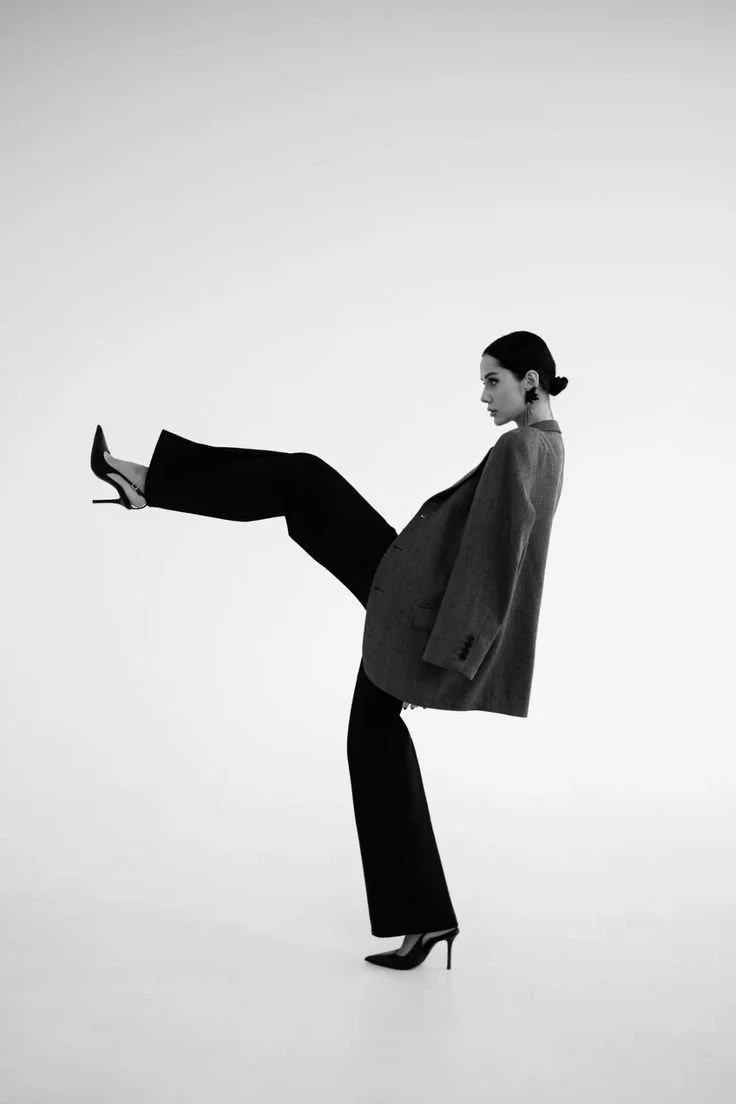 A woman in black heels, a blazer, and black pants balancing on one leg with the other leg extended forward.