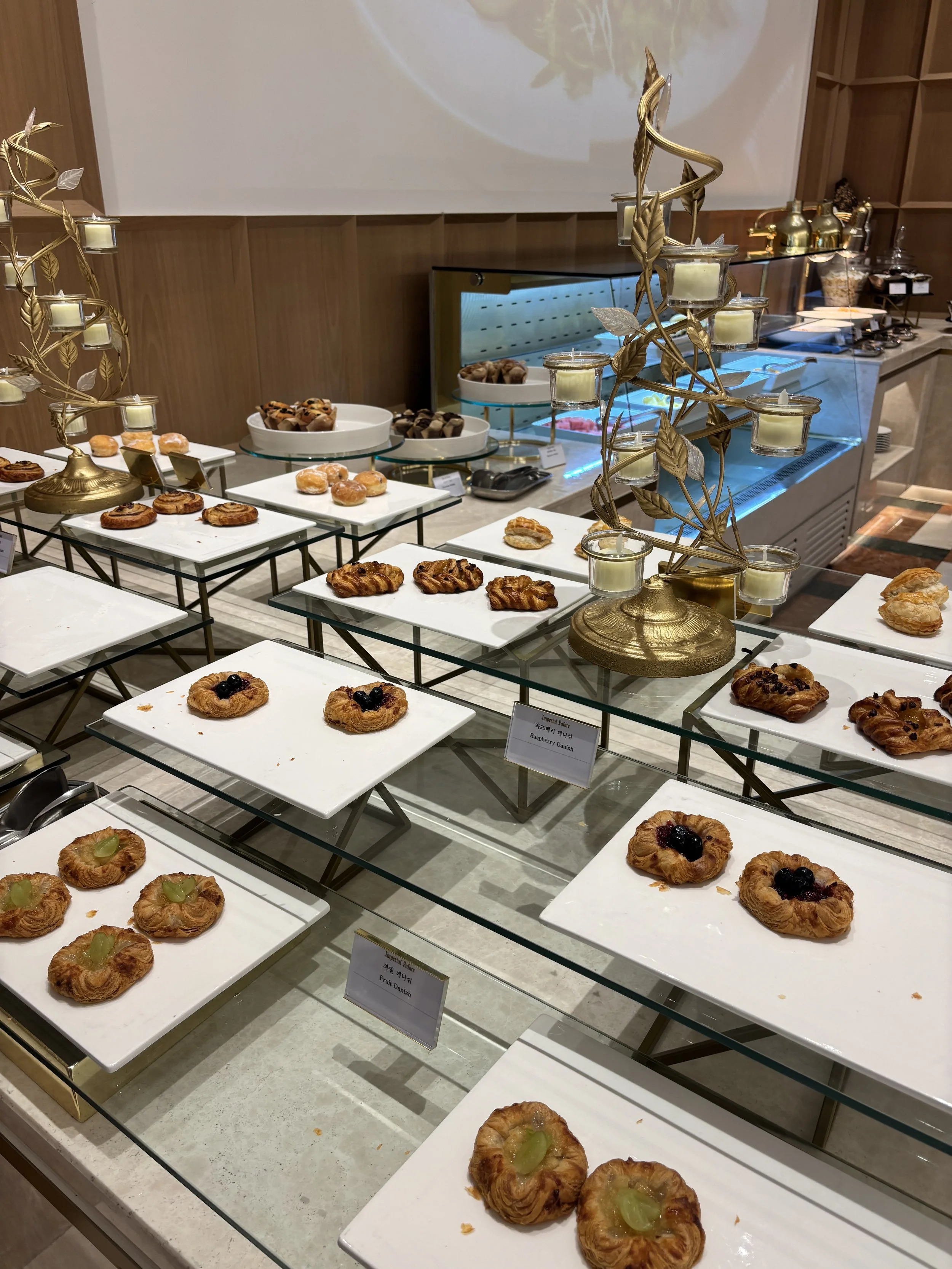 A dessert display with various pastries including fruit Danish, cream Danish, and other sweet treats on white rectangular and circular plates, with labels indicating the types of pastries, in a buffet setting with a wooden wall and glass countertop.