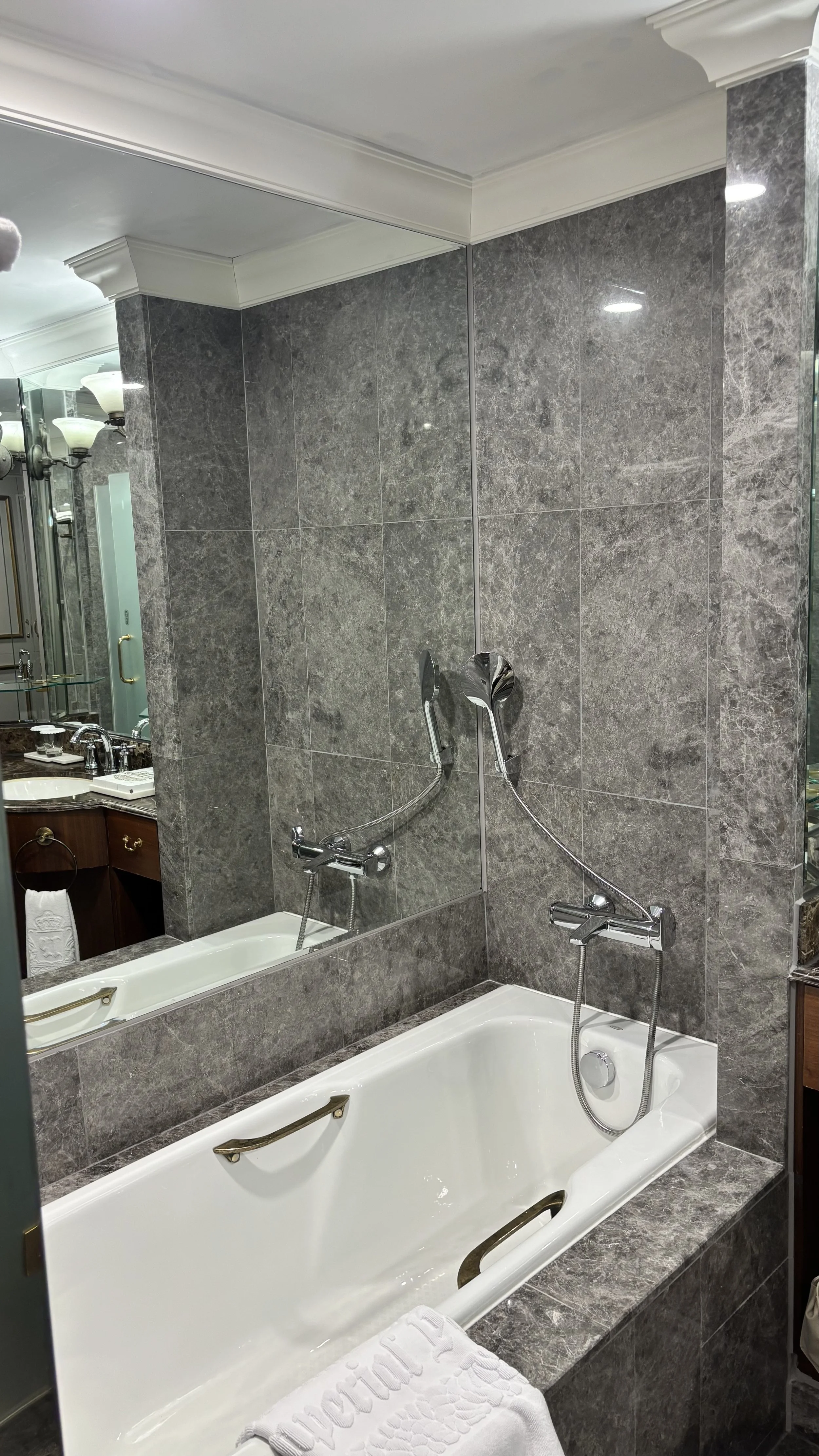 A bathroom with a bathtub and grey tiled walls, featuring a large mirror, a towel at the end of the bathtub, and a double shower head with handheld attachments.