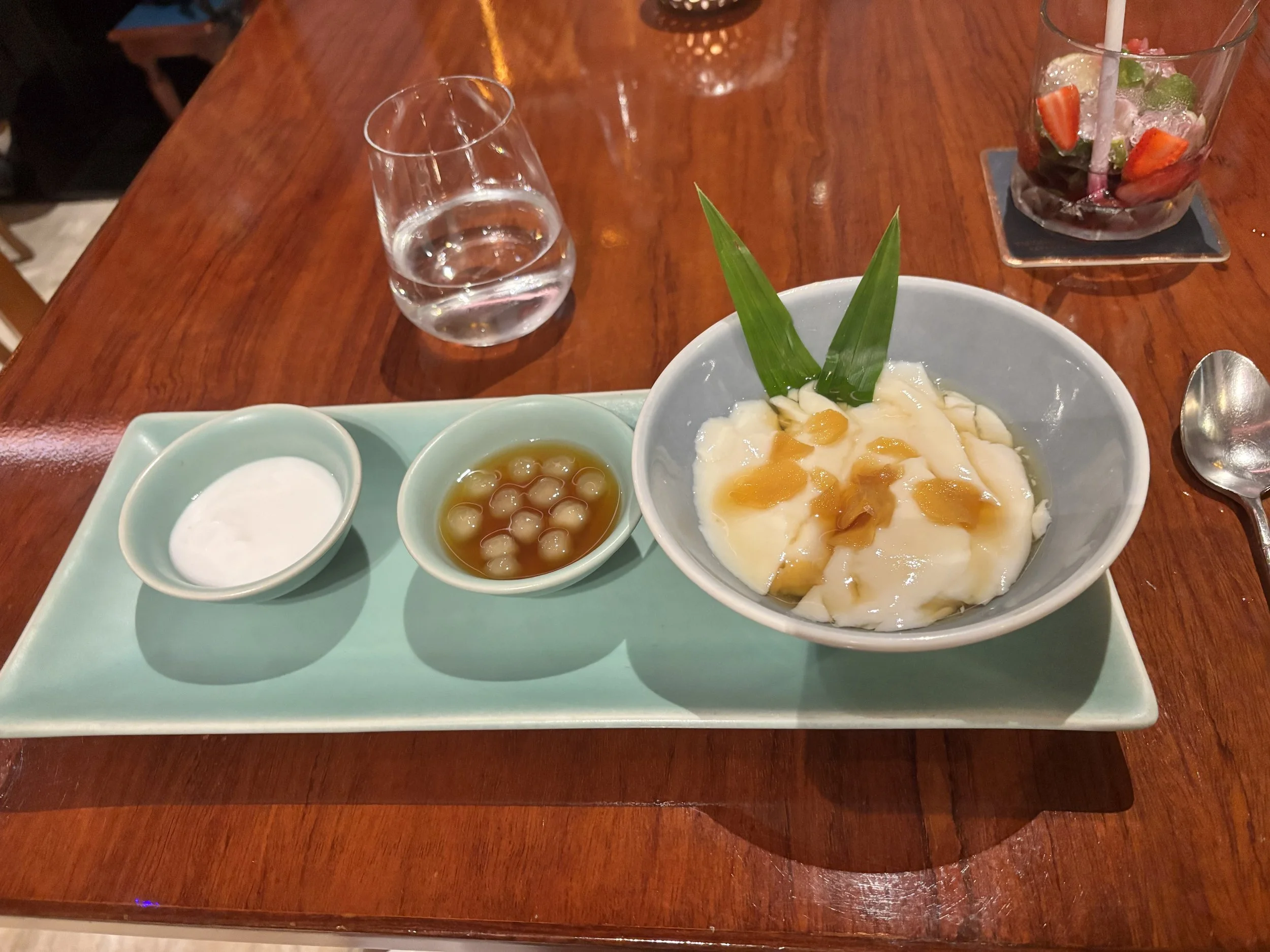 A wooden table with a glass of water, a small bowl of tapioca pearls in syrup, a bowl of creamy dessert topped with yellow fruit pieces and a green leaf, dipping sauces, and a glass of fruit-infused water with strawberries and mint.