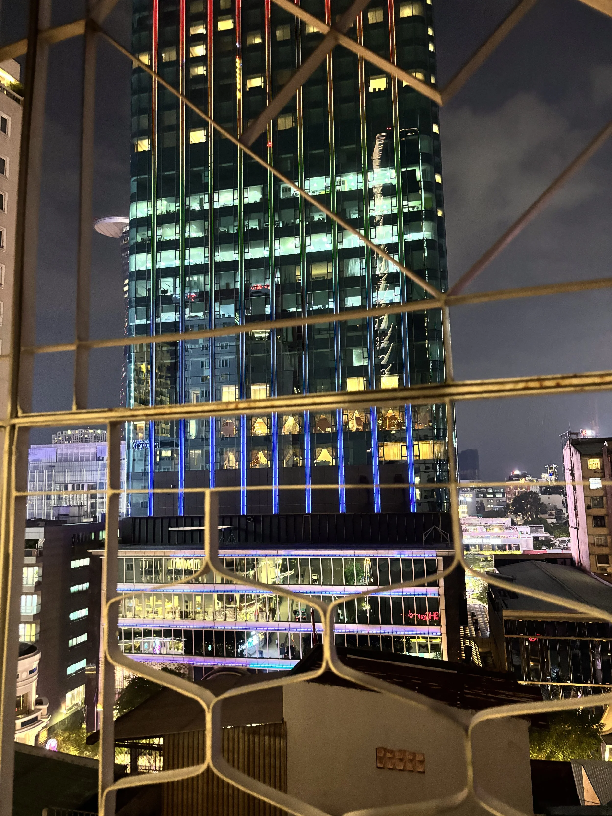 Nighttime cityscape view through a wire fence, featuring a tall glass building illuminated with green, blue, and yellow lights, with other illuminated buildings visible in the background.