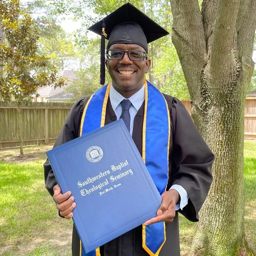 A man in graduation attire holding a diploma certificate outside near a tree, with a fence and trees in the background.