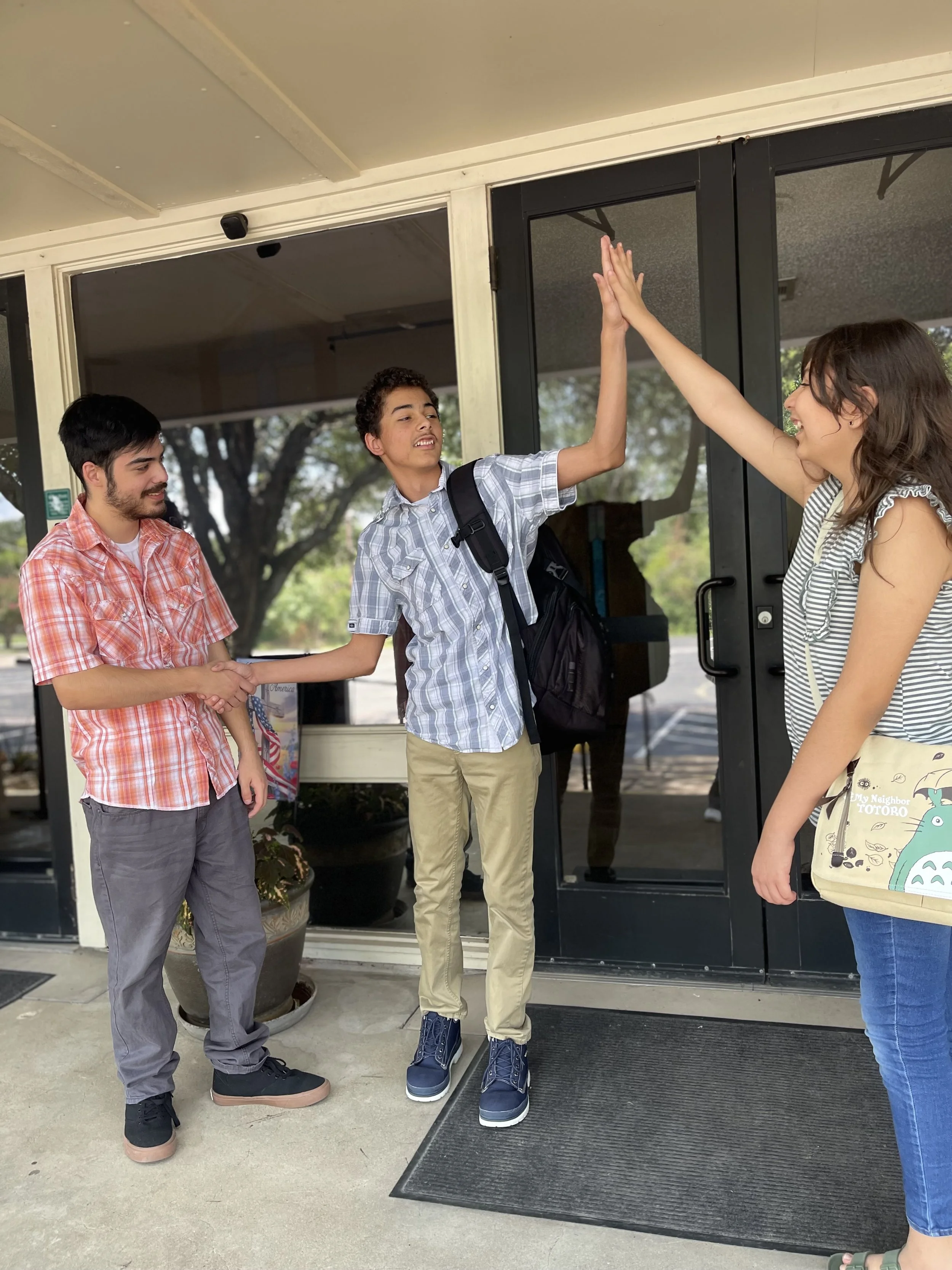 Three young people standing outside a building entrance, high-fiving each other, with one person wearing a striped shirt and another wearing a backpack